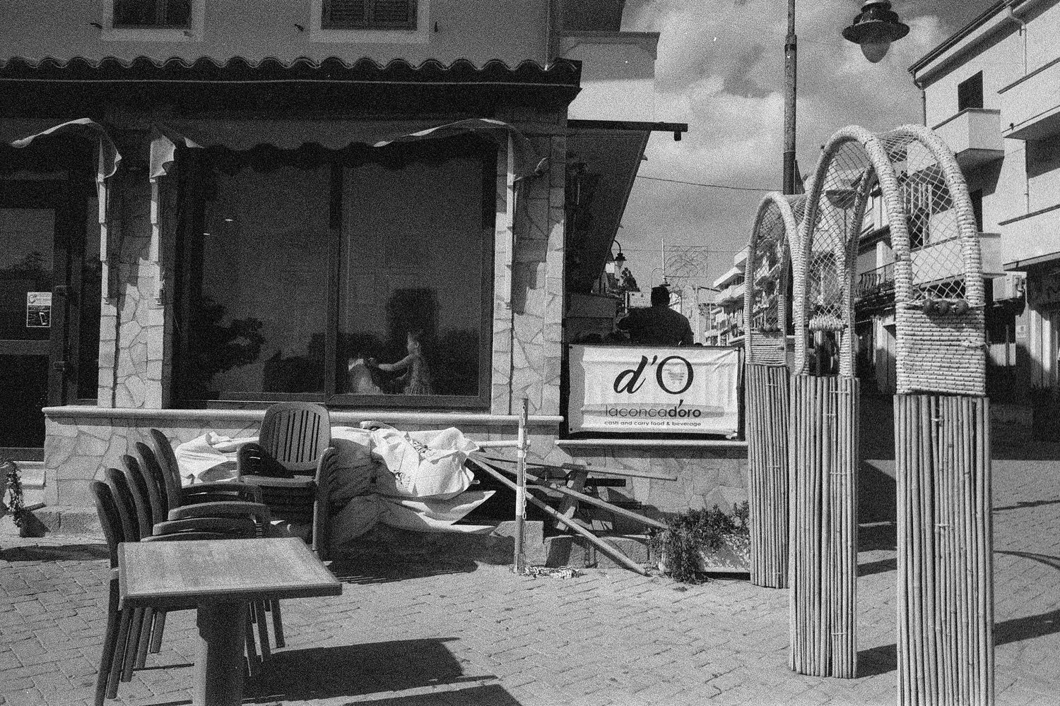 Black and white vintage photo showing empty patio chairs at a covered outdoor dining area with arched architectural details.