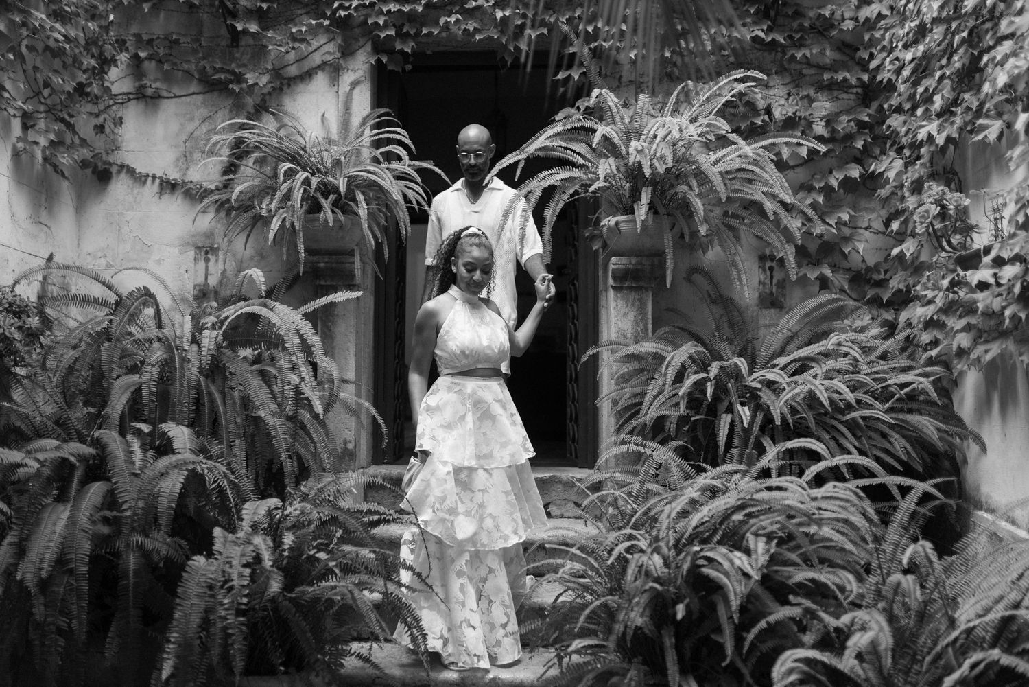 A black and white wedding photography scene in a garden courtyard surrounded by lush tropical plants and ferns.