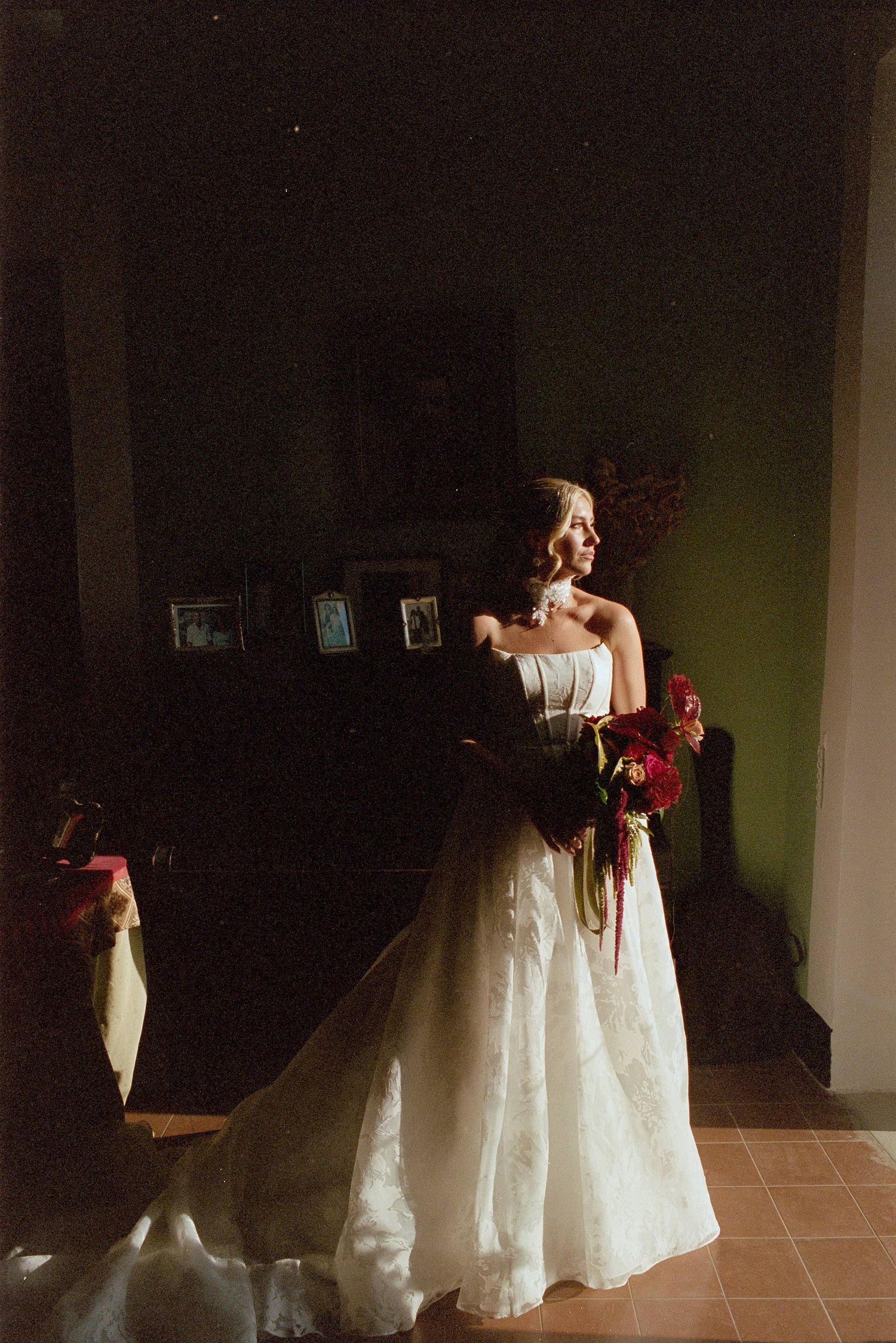 A person in a white wedding gown holds red flowers while standing in dramatic side lighting near framed photos.