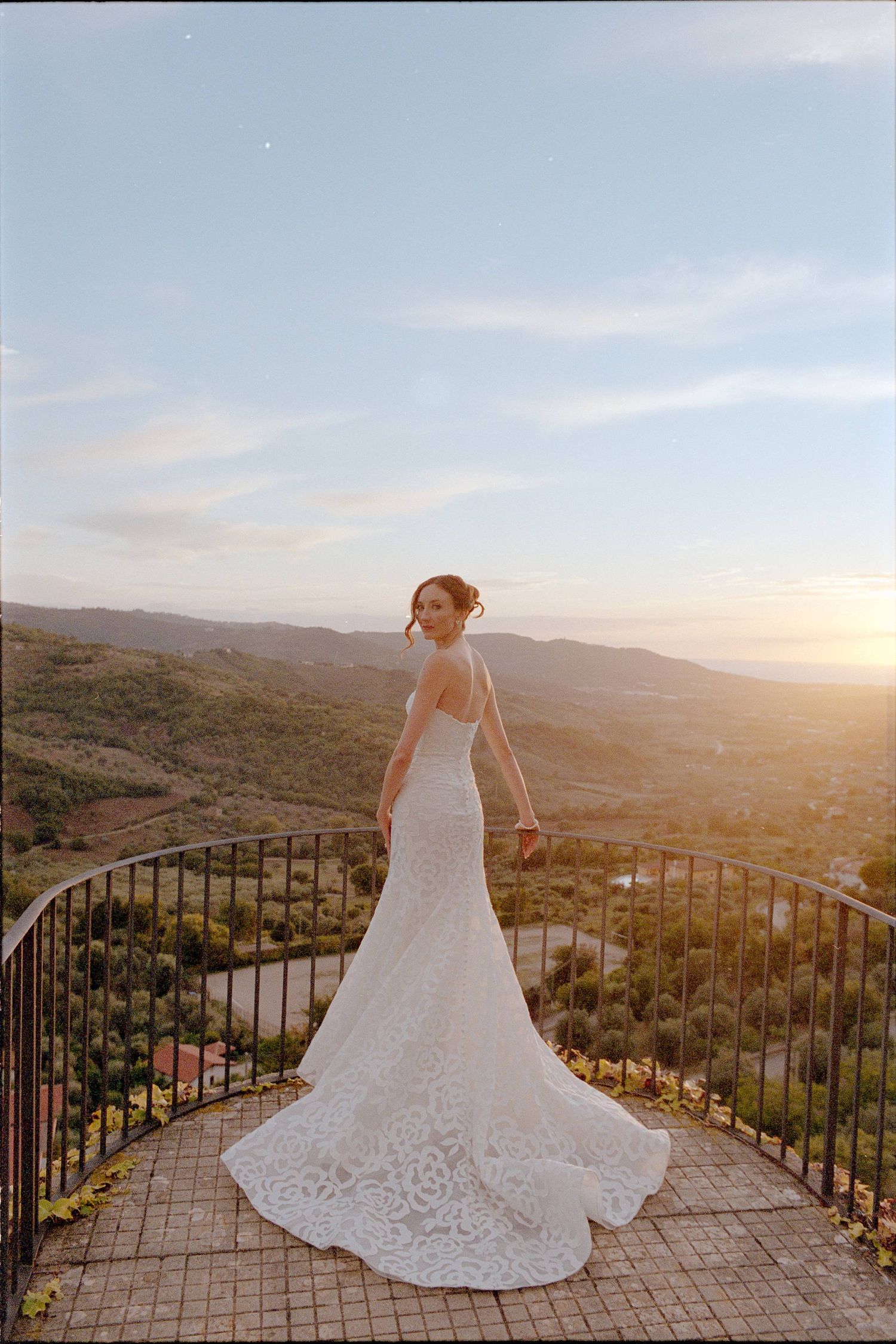 A bride in a flowing lace wedding dress poses on a scenic overlook at sunset with mountain views in the background.