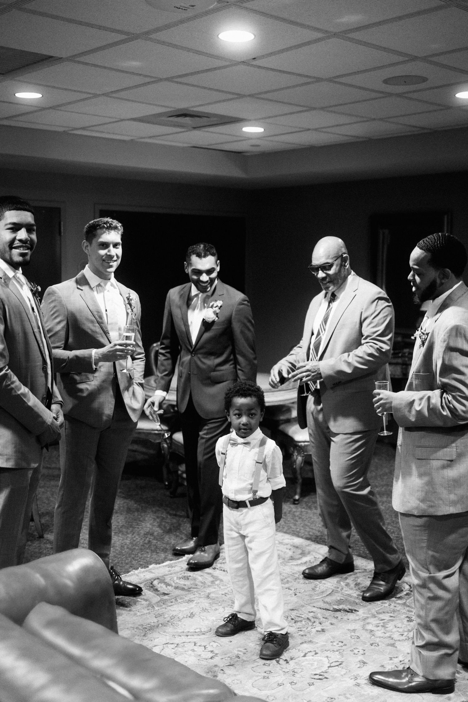A small child in formal attire stands surrounded by groomsmen in suits at what appears to be a wedding gathering.