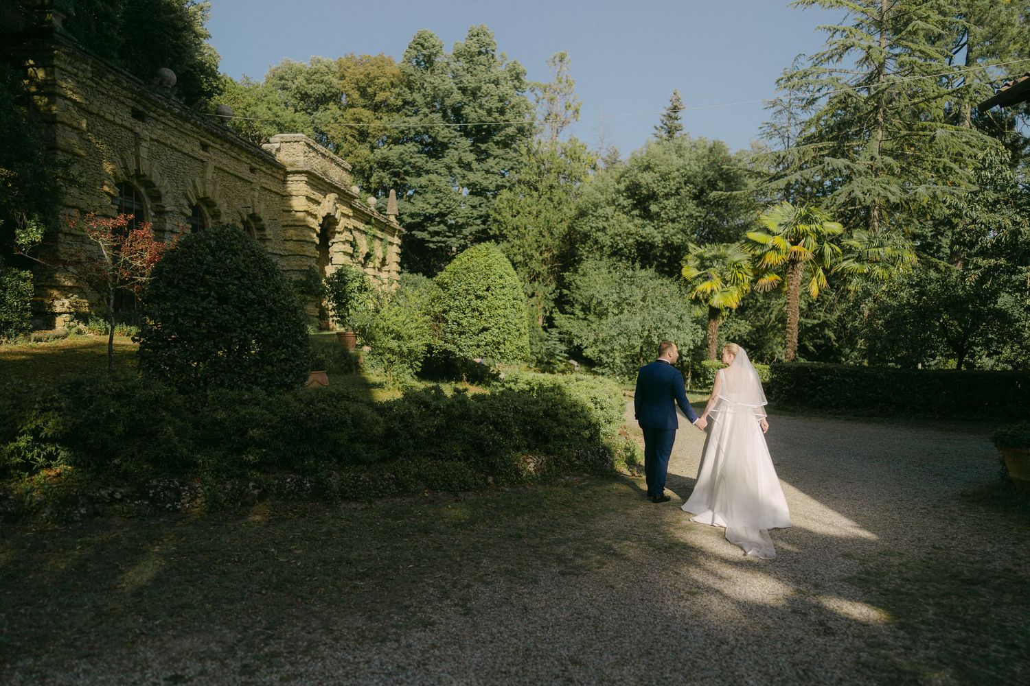 A couple in wedding attire walks along a tree-lined garden path on their special day.
