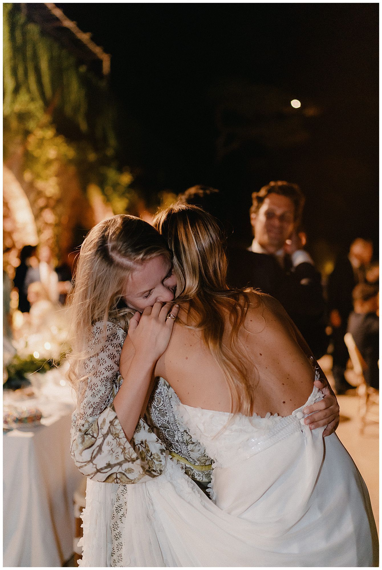 Two people share an emotional embrace at a dimly lit outdoor wedding reception with candles and decorative lighting.