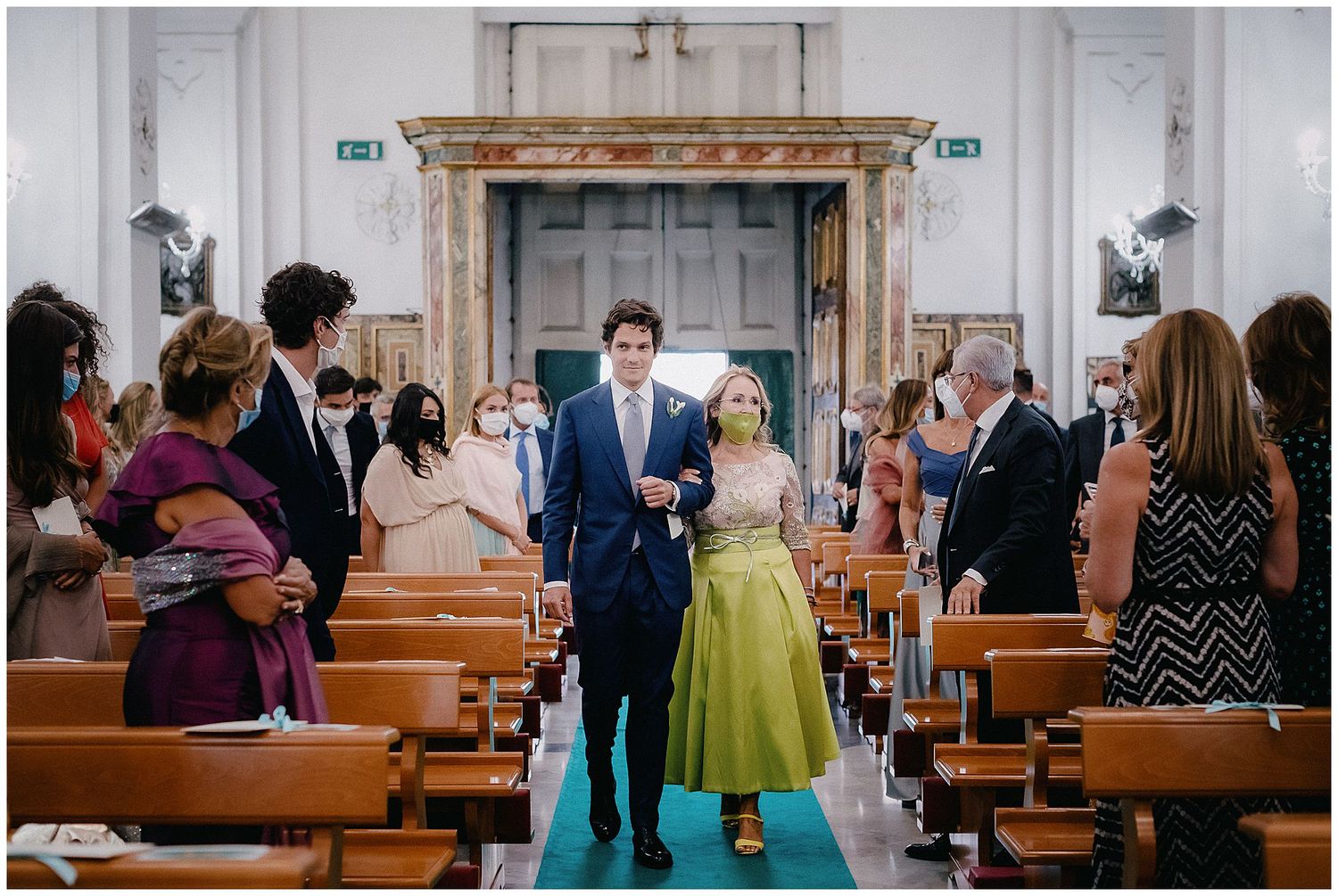 Wedding guests stand in church pews as couple walks down aisle lined with turquoise carpet during ceremony.