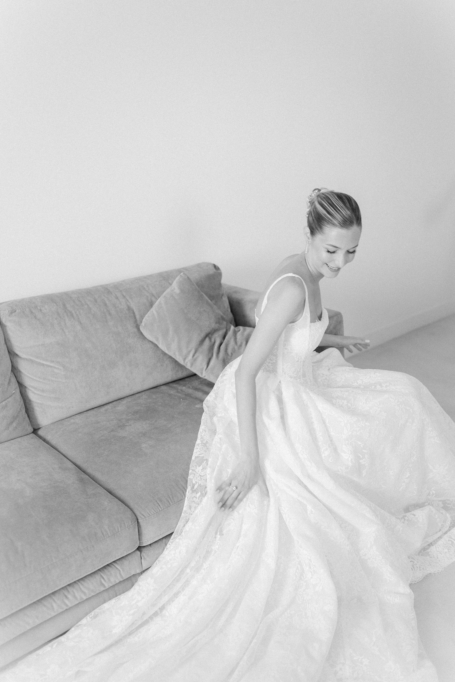 Black and white photo of a smiling bride in a lace wedding gown arranging her dress train beside a gray sofa.