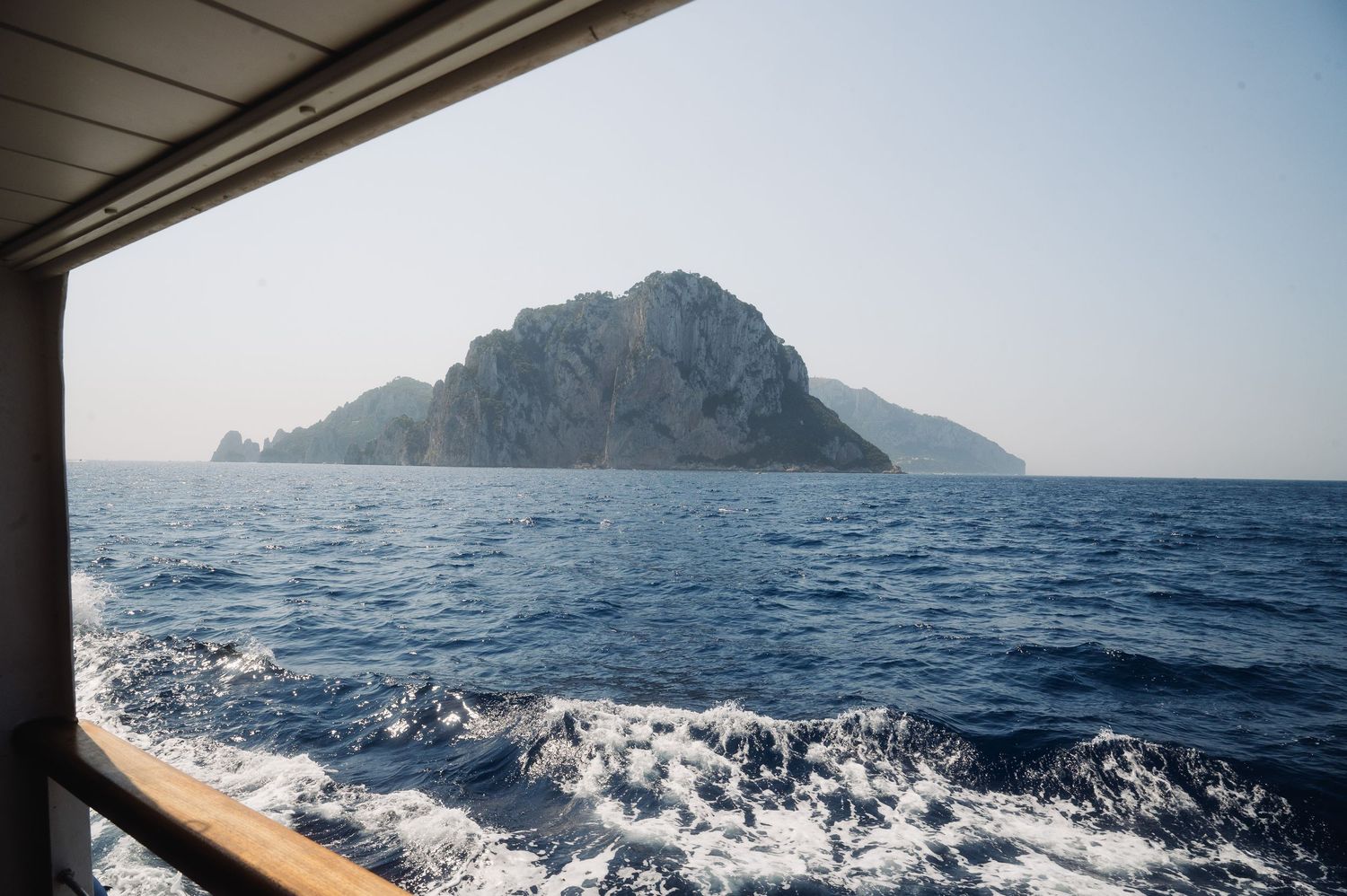 Rocky island rises from blue Mediterranean waters viewed through boat window with white wake visible below.