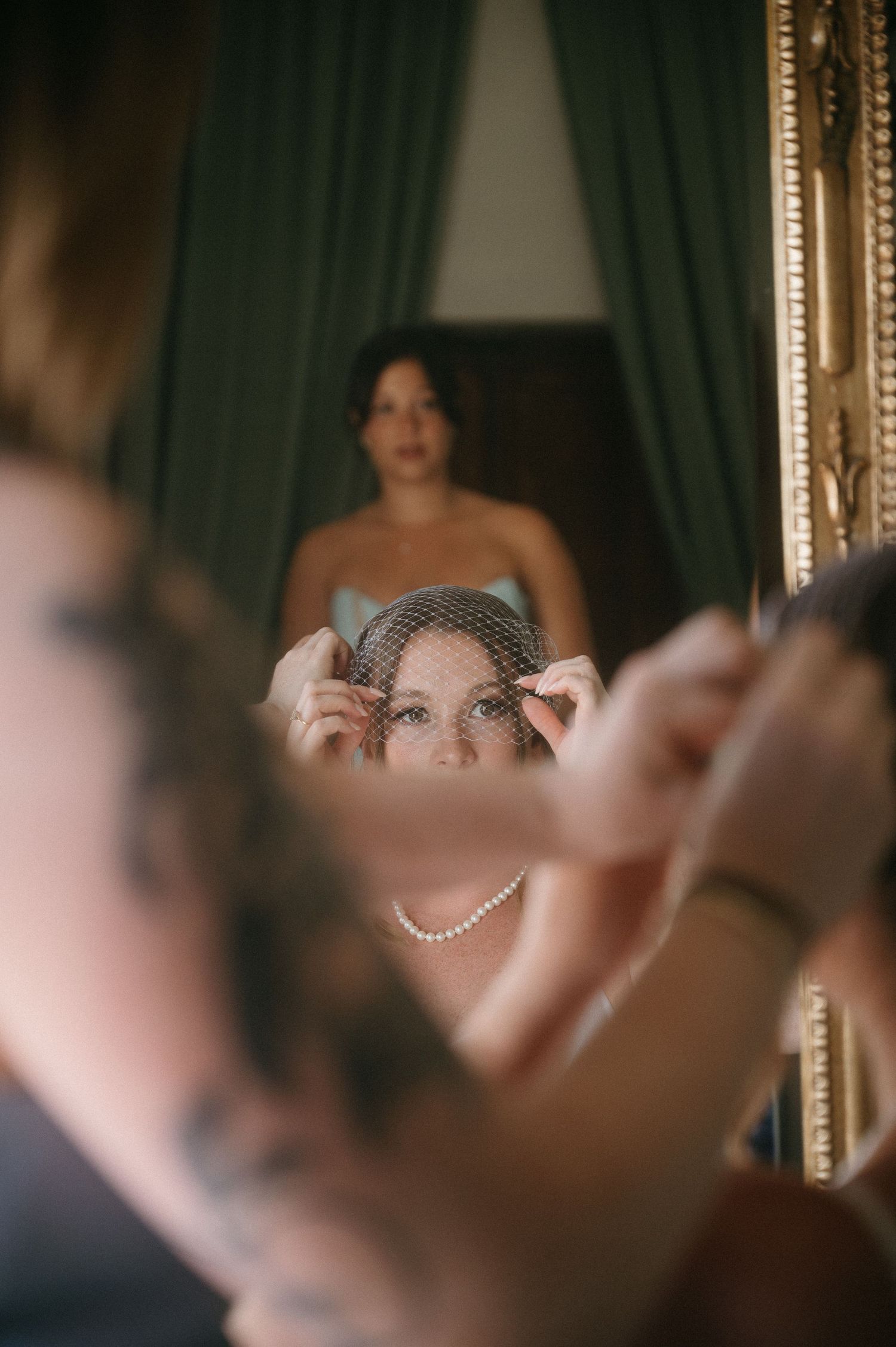 Reflection in ornate mirror shows delicate adjustments being made to a bridal veil with green curtains in background.