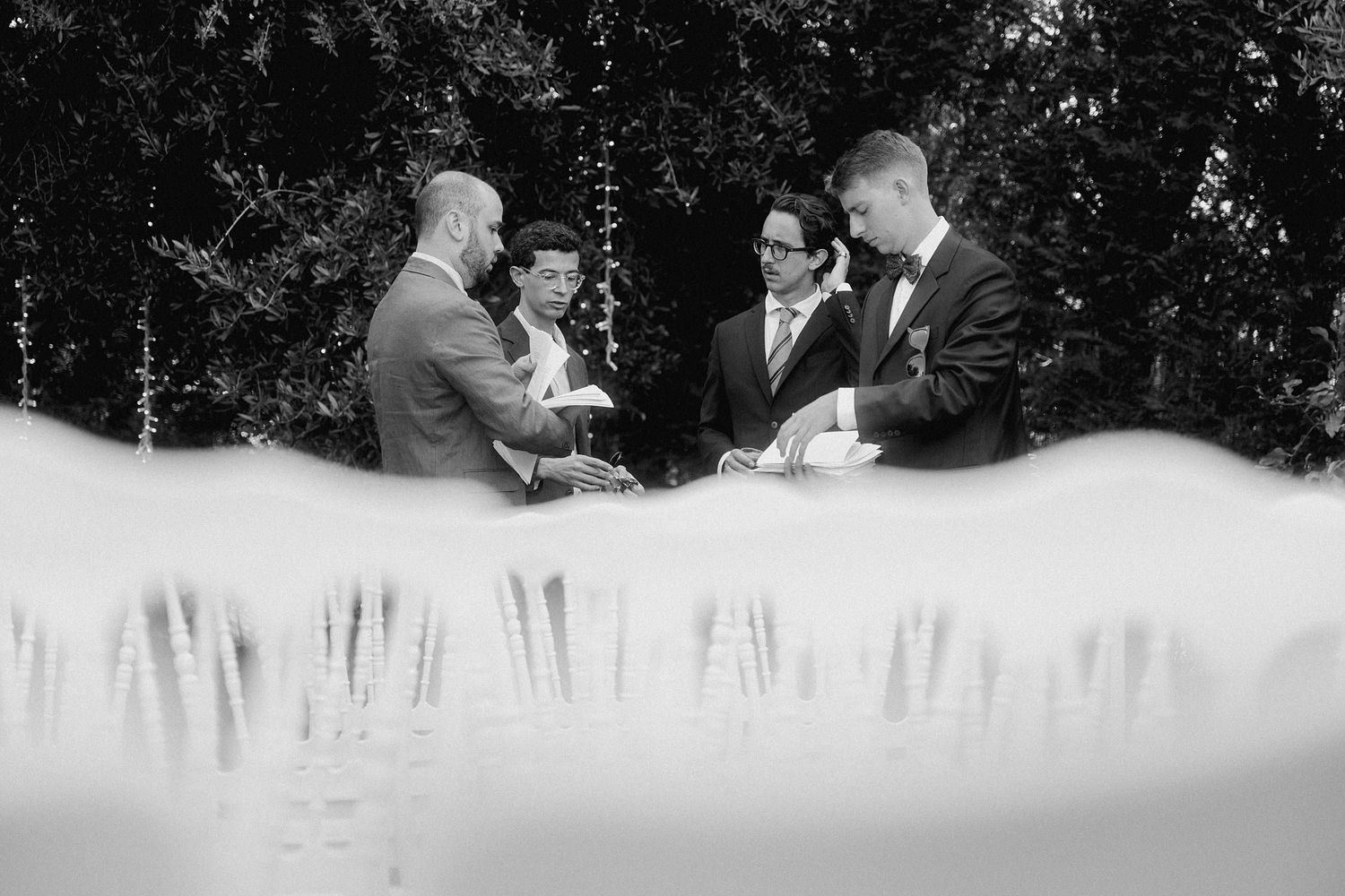 A black and white candid photo of people in formal attire having a conversation outdoors, framed by blurred foliage.