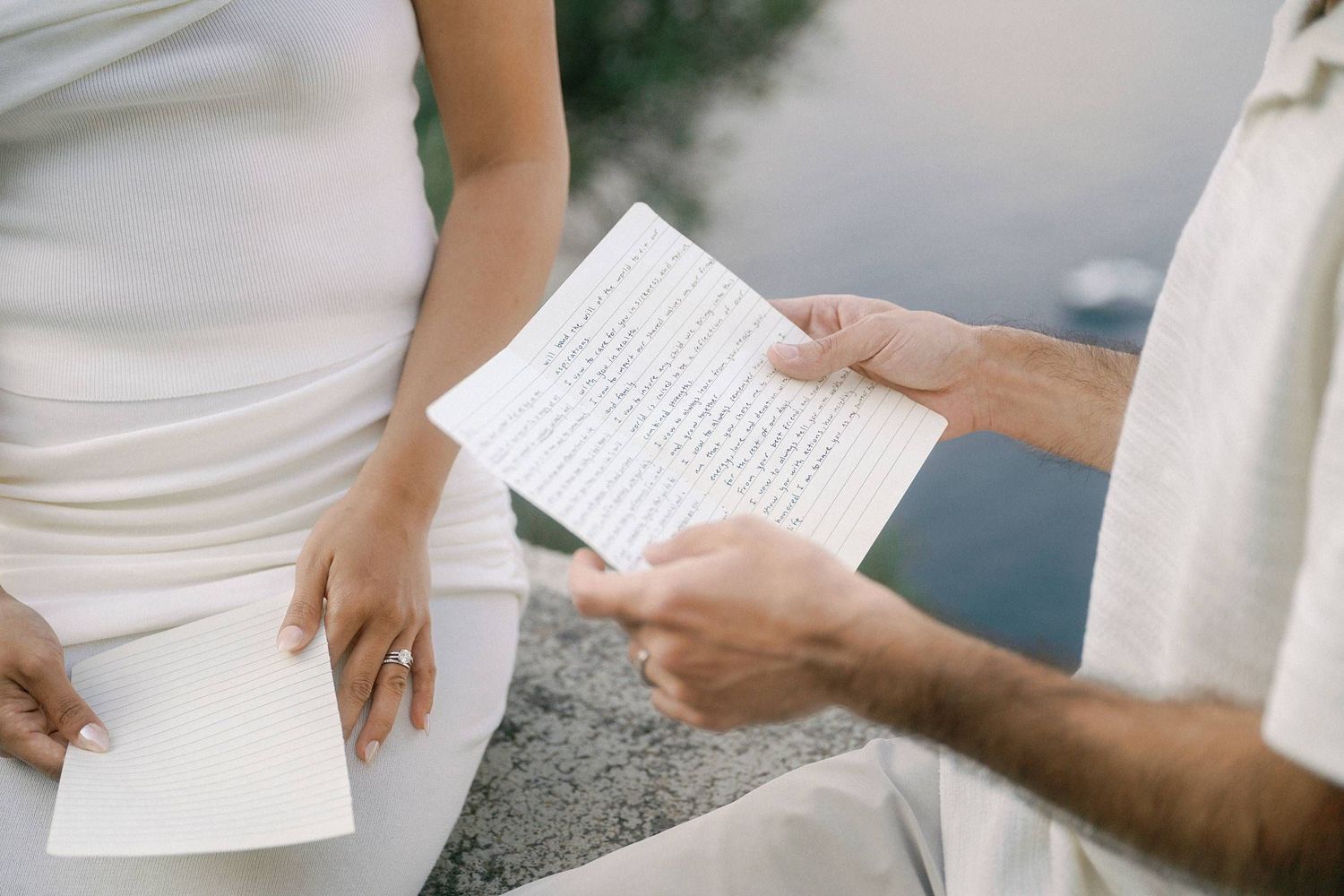Two individuals in white clothing hold handwritten vows or letters while seated by the ocean.