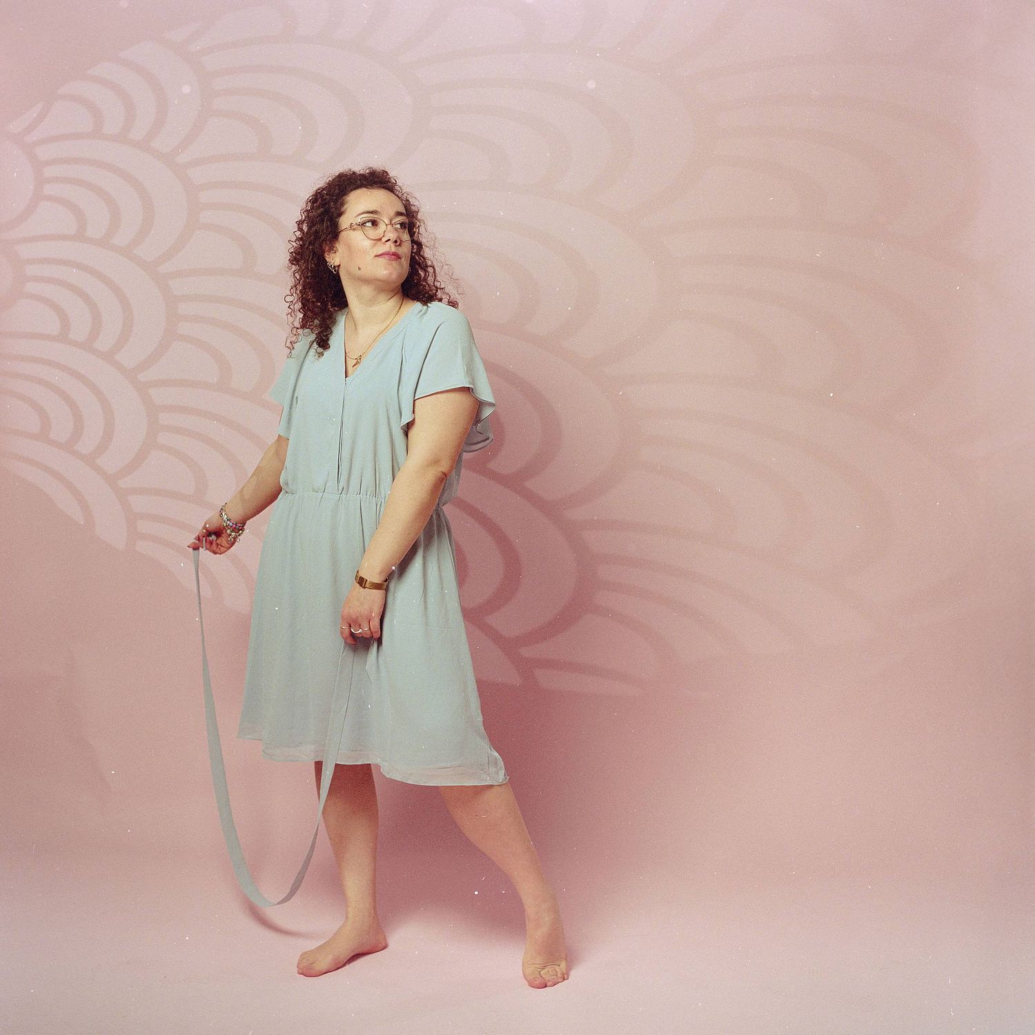A person in a mint green dress stands barefoot against a pink backdrop with decorative swirl patterns.