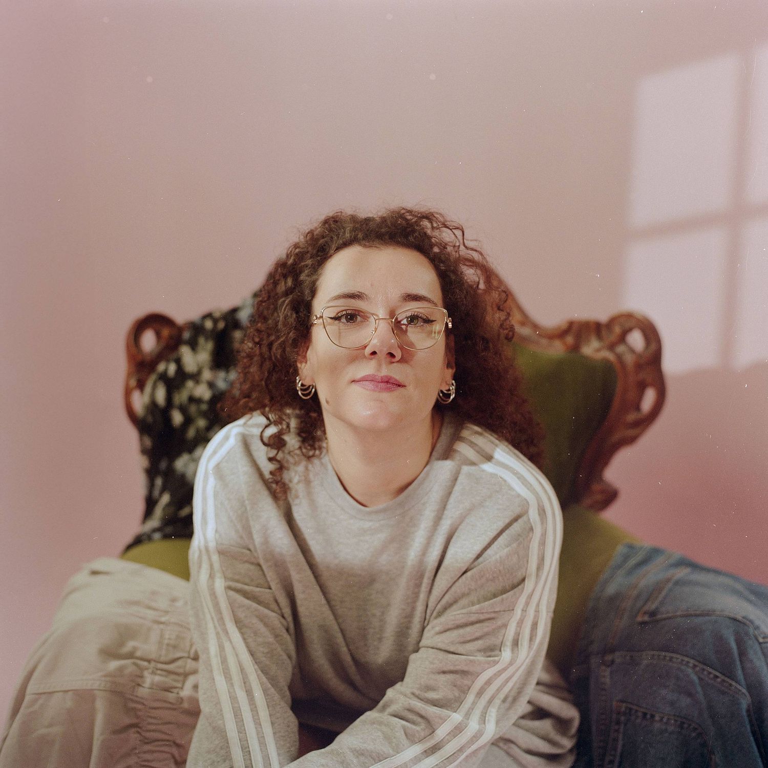 Someone with curly hair and glasses relaxes on a vintage green chair in a softly lit pink room.