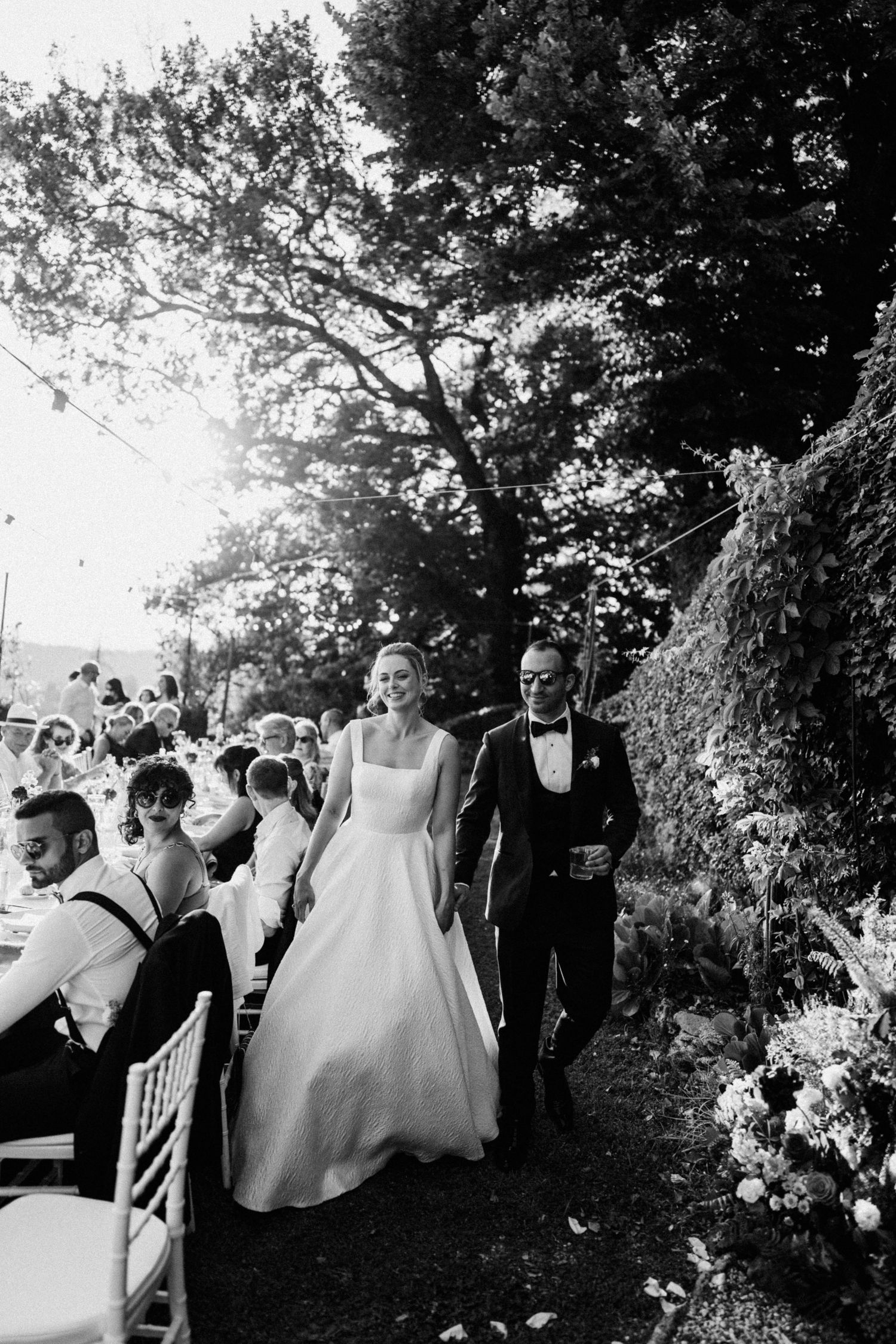 A black and white wedding photo showing guests seated at an outdoor ceremony with a couple walking down a garden path.