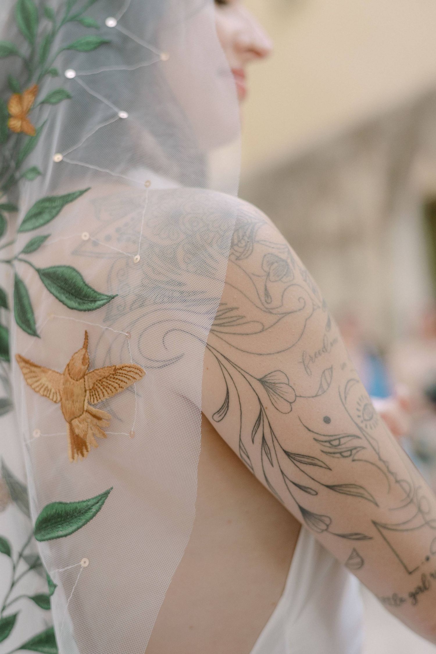 Close-up of a bride's shoulder showing delicate floral tattoos with a golden hummingbird design through sheer veil fabric.