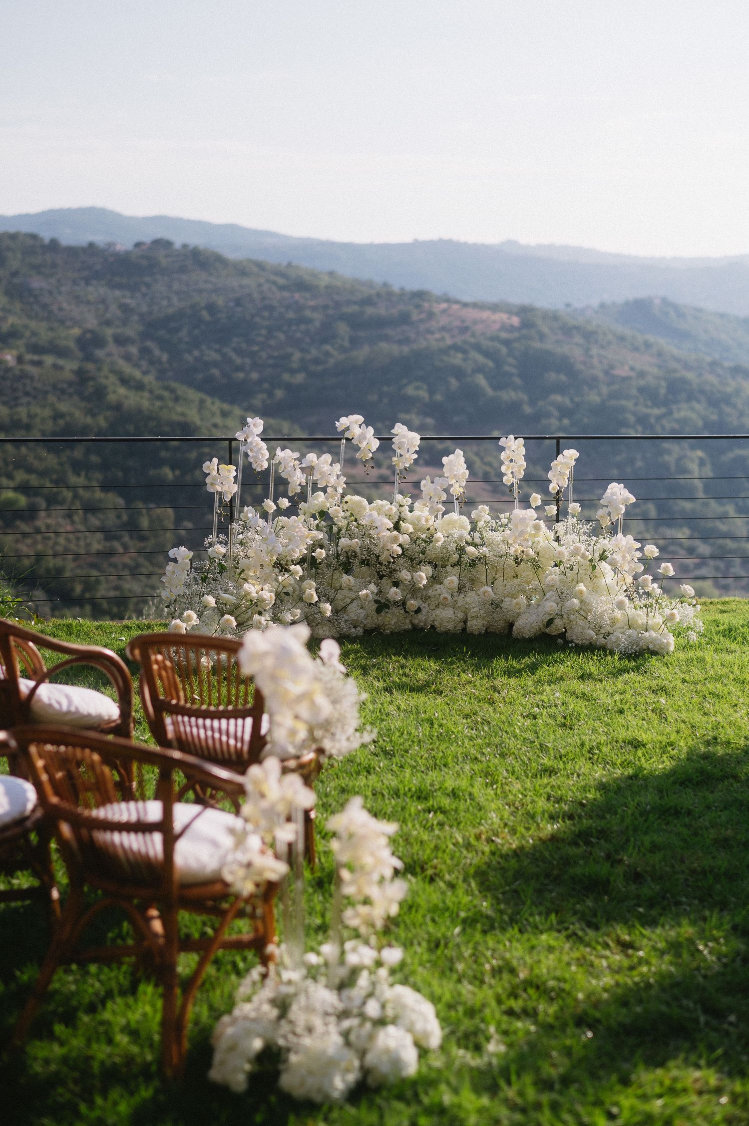 Rattan chairs and white floral arrangements set against rolling hills create an intimate outdoor wedding ceremony setting.