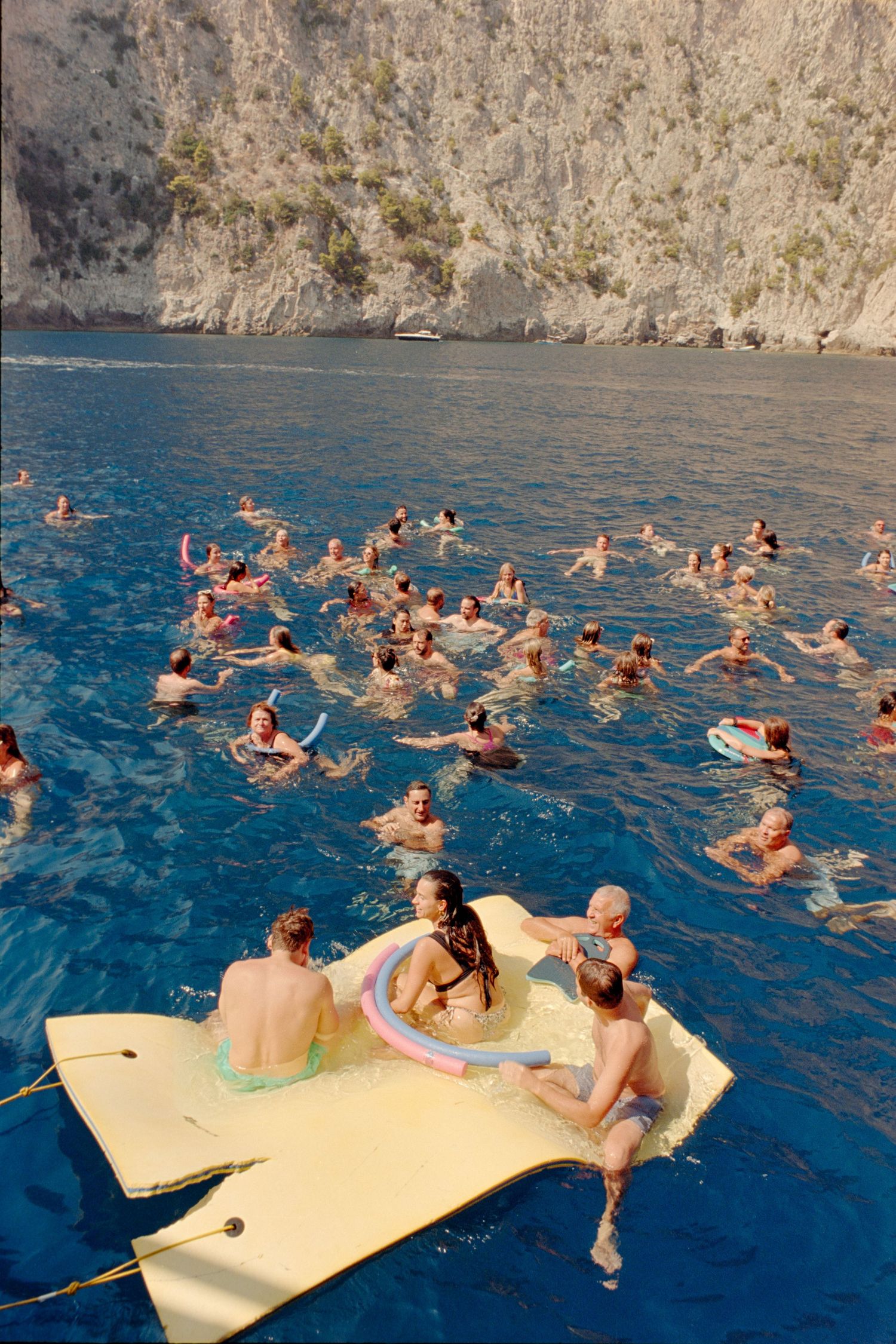 A group of swimmers float in blue Mediterranean waters near a rocky cliff while relaxing on a yellow floating mat.
