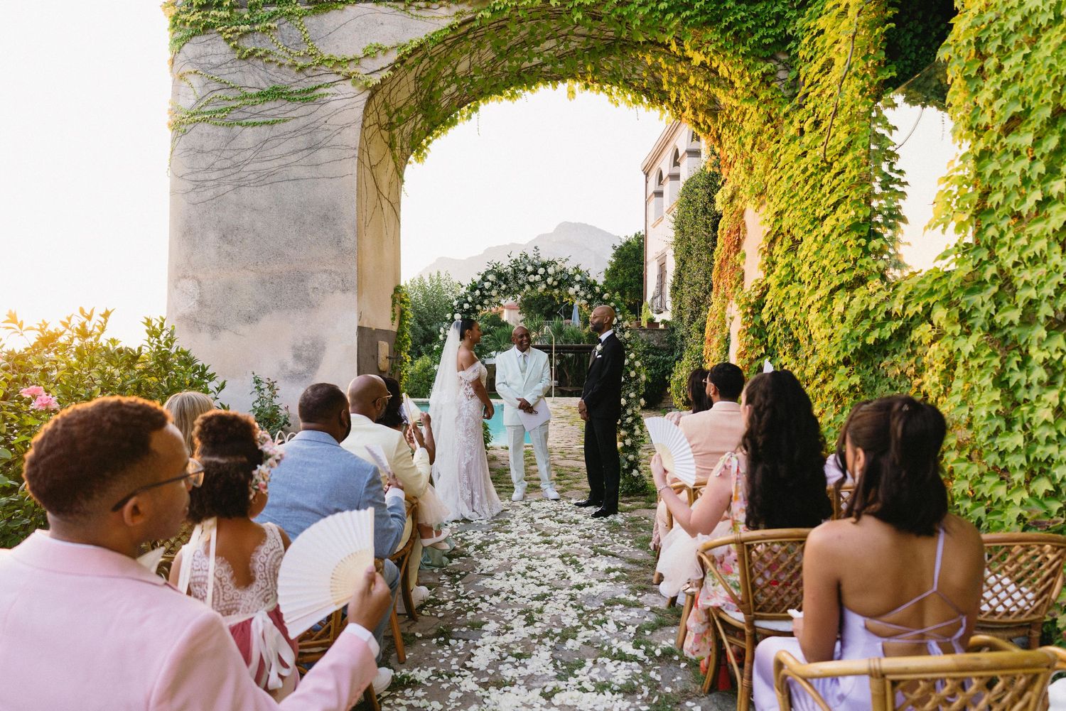 Wedding guests gather under a vine-covered pergola during an outdoor celebration at a scenic venue.