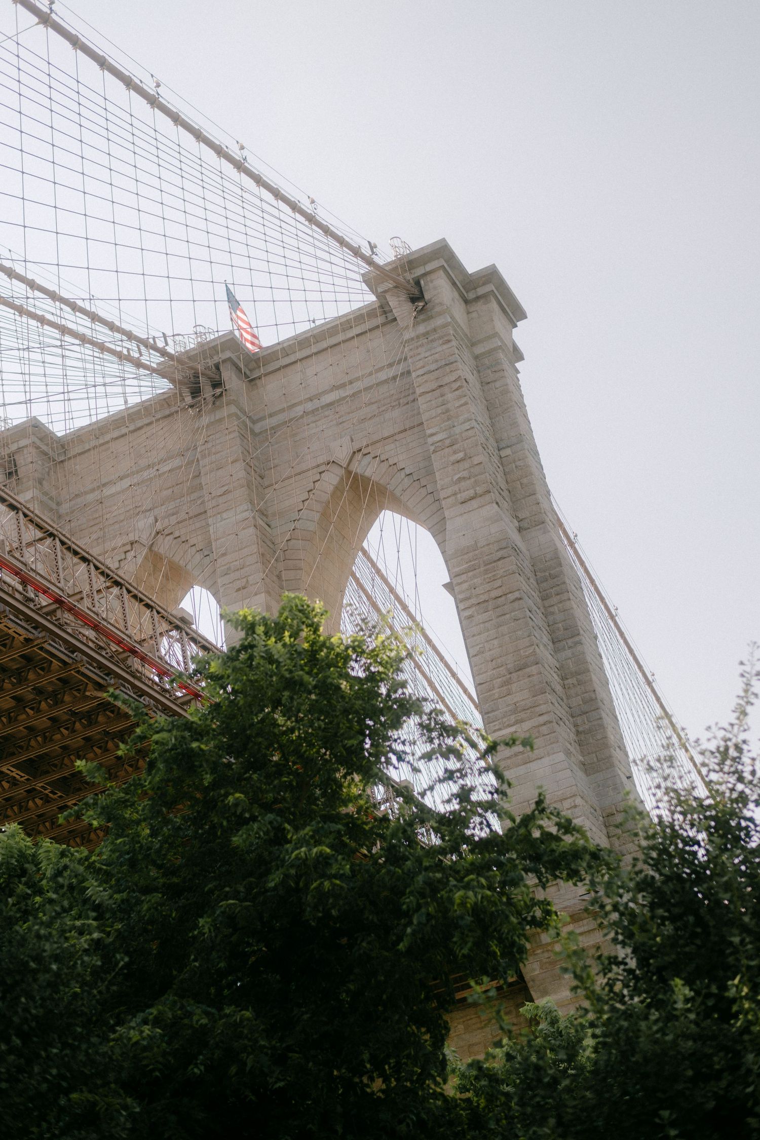 Low angle view of the Brooklyn Bridge tower with American flag and green trees in the foreground against cloudy sky.