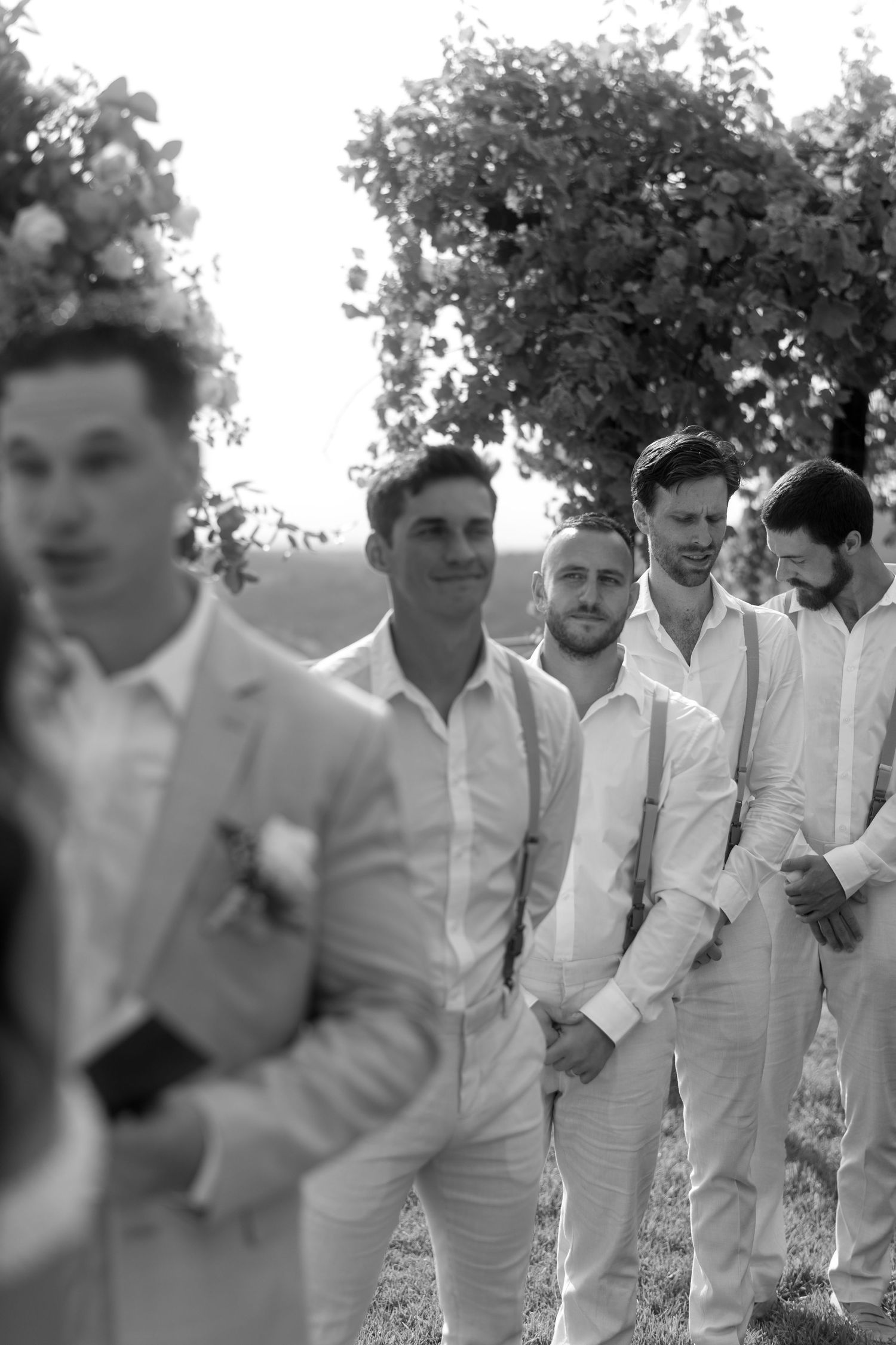 A line of groomsmen in white suits stand together outdoors in a candid black and white photo.