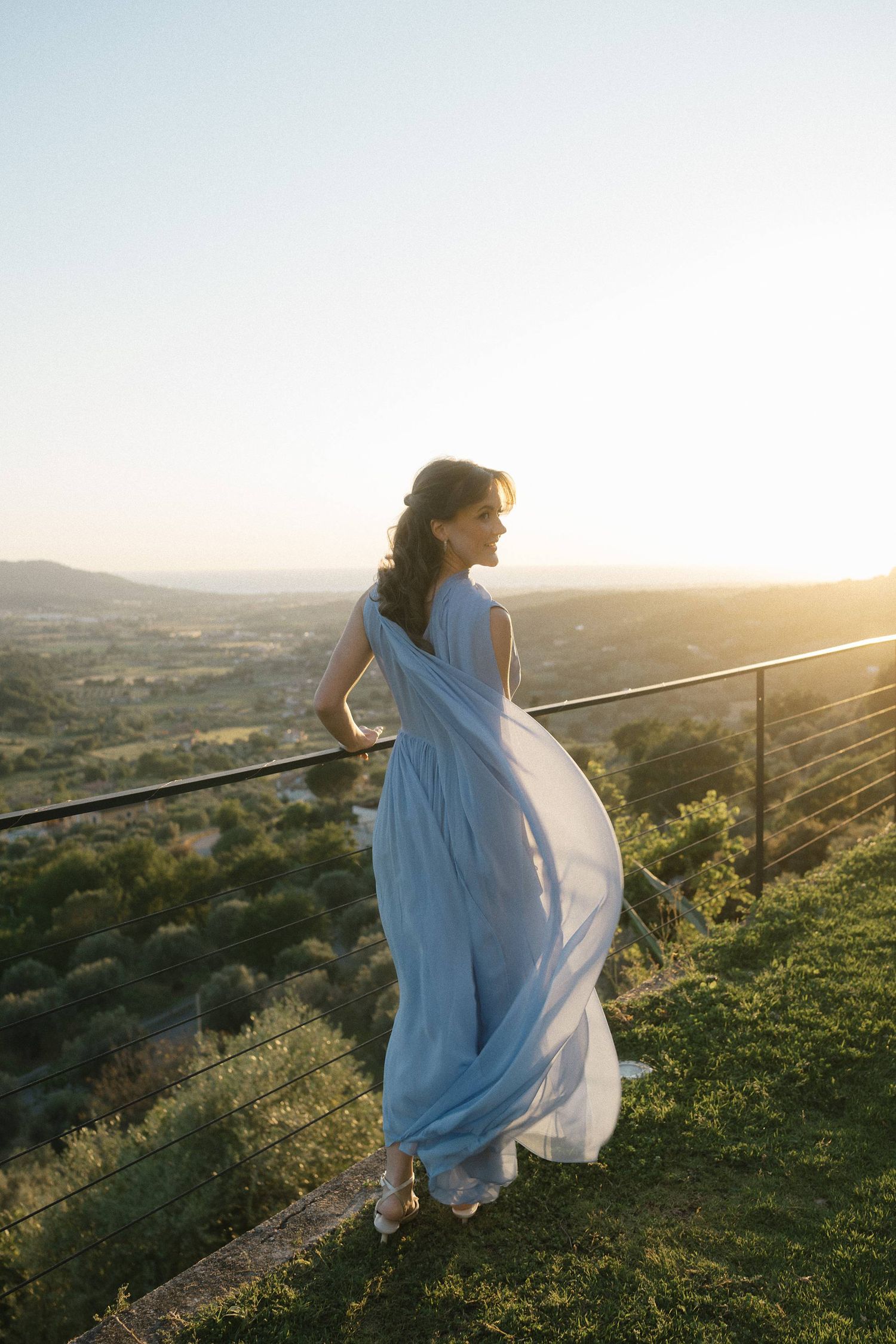 Woman in flowing blue gown stands on scenic overlook at golden sunset with rolling hills in background.