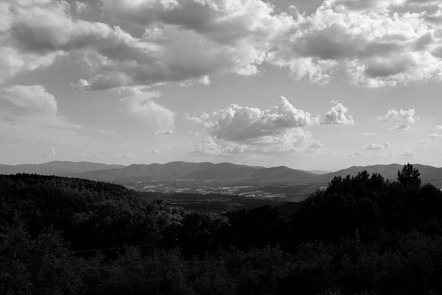 Black and white landscape photo of rolling mountains with dense forests under dramatic cloudy skies.