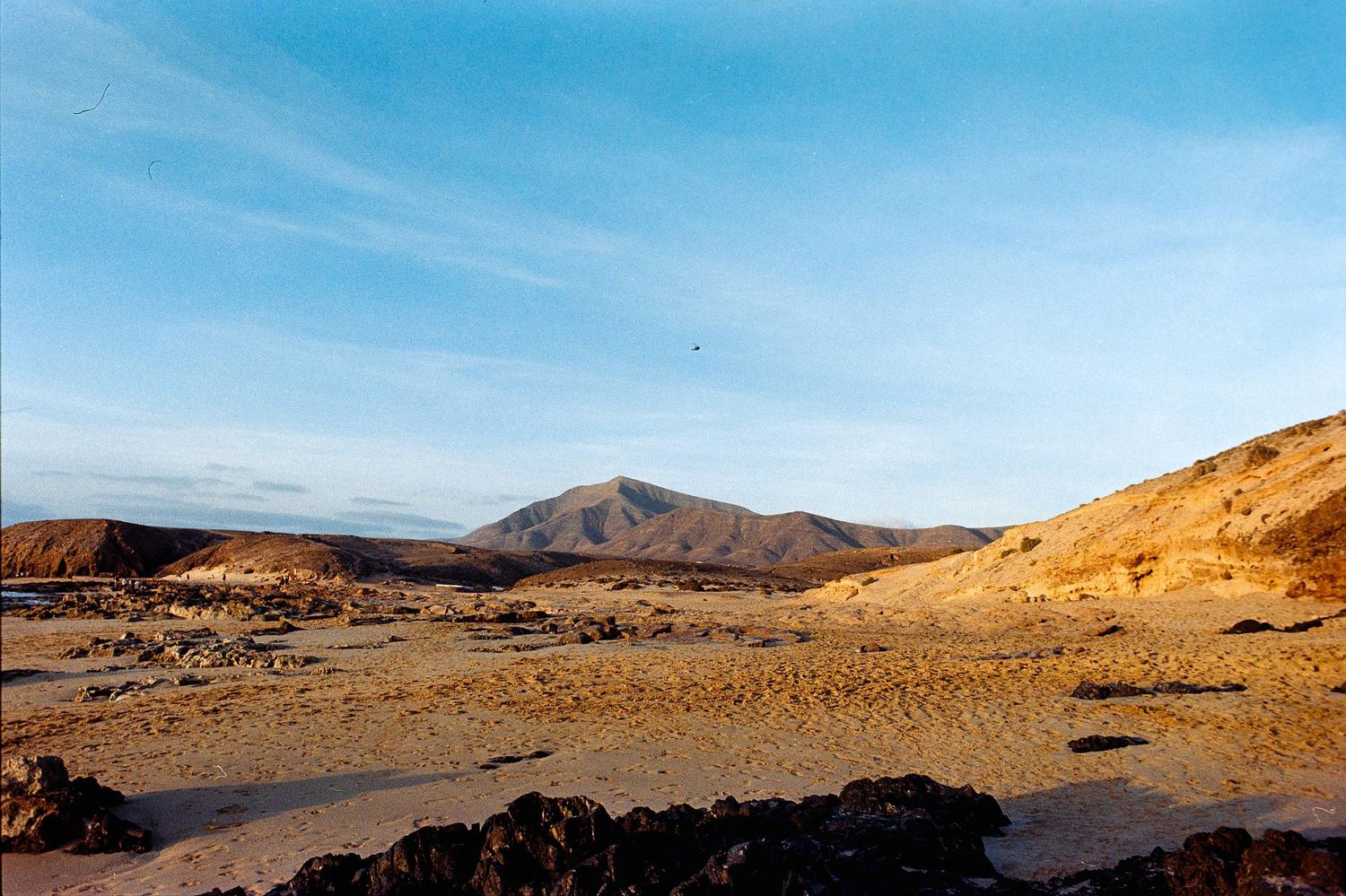 Arid desert landscape with golden sand dunes and distant mountains under bright blue sky and wispy clouds.