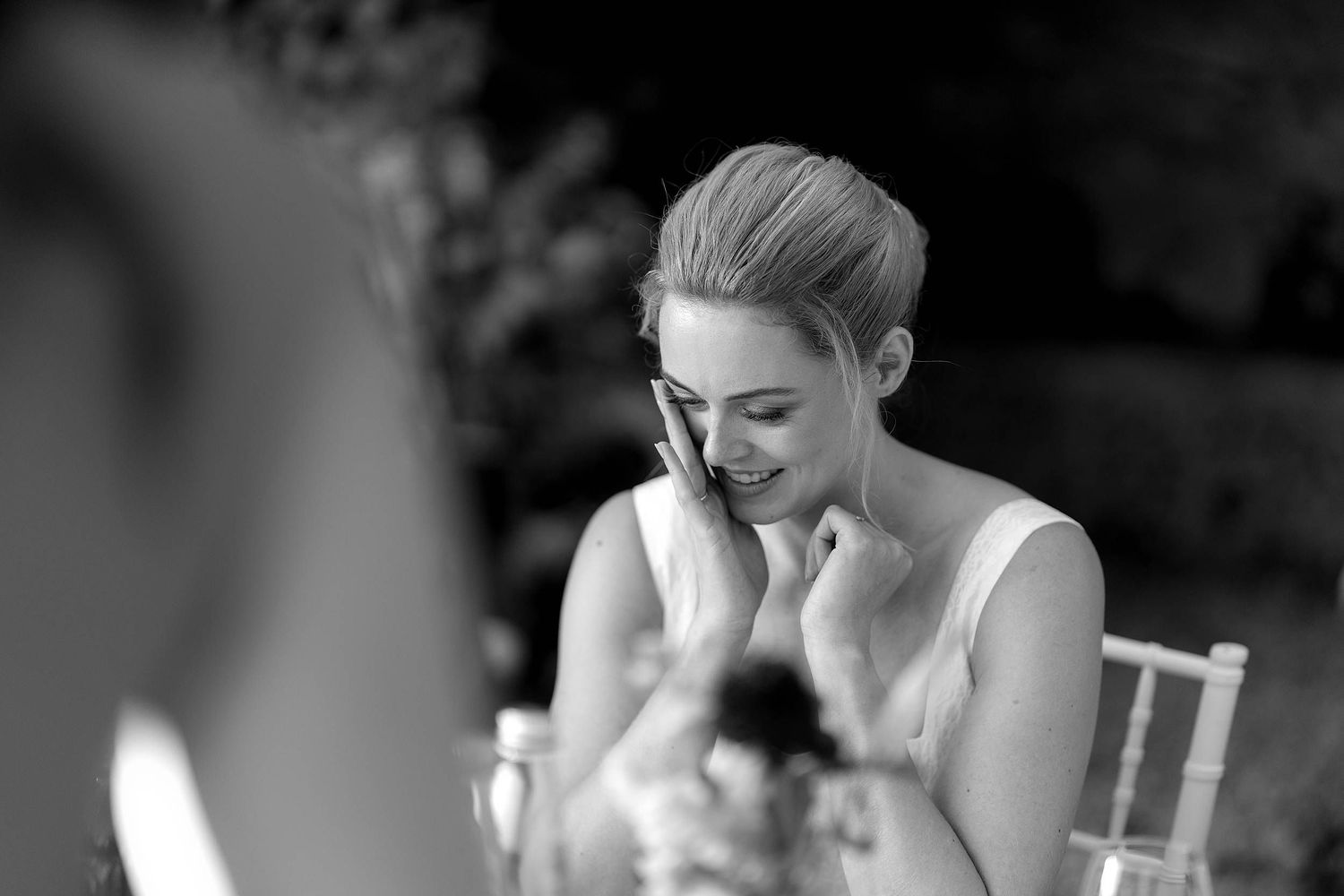 Black and white candid photo of someone in white attire seated at a table showing emotional expression during conversation.