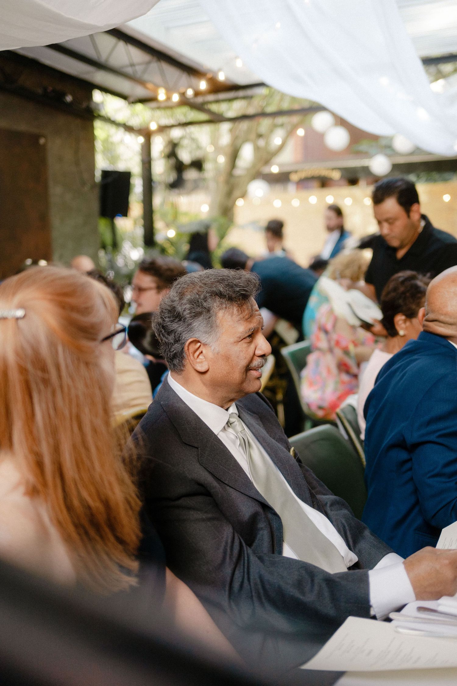 Guests gather at an outdoor evening event with string lights overhead and people seated at tables.
