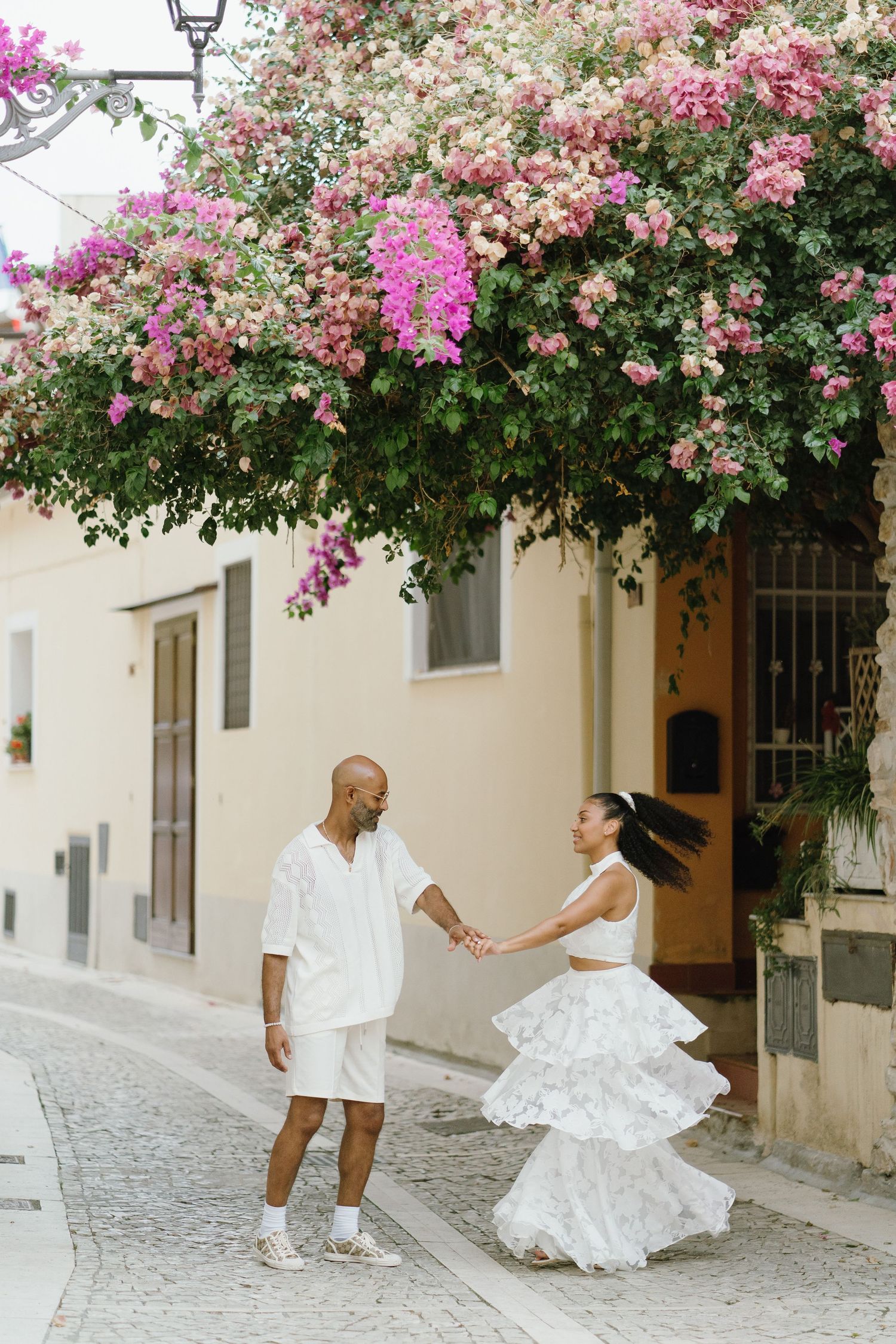 A couple dances in a charming cobblestone street under blooming pink bougainvillea flowers in a Mediterranean setting.