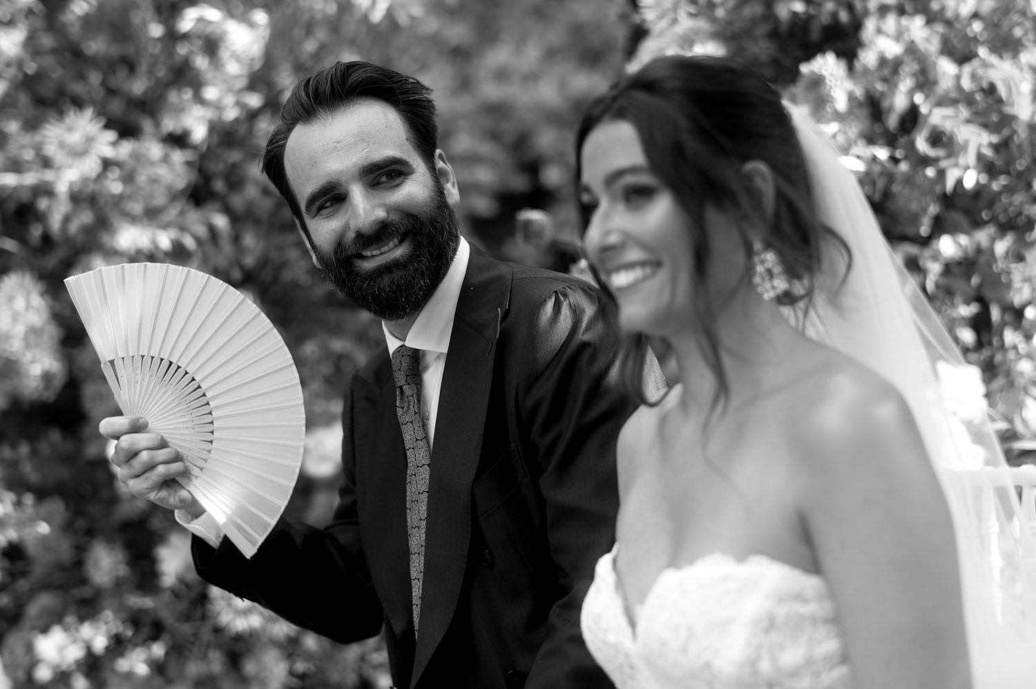 A black and white wedding photo shows a couple outdoors, with the bride holding a decorative hand fan.