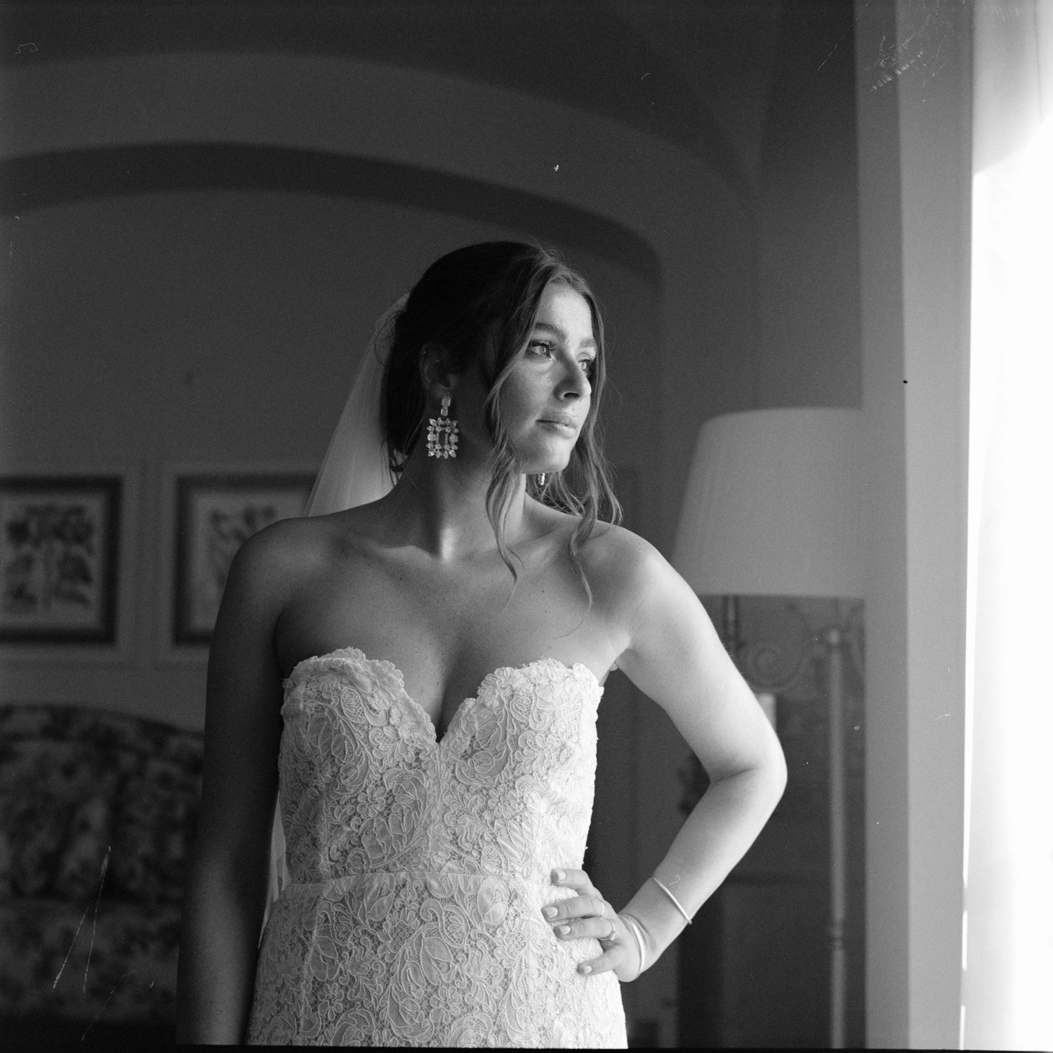 Black and white photograph of a bride in a lace wedding dress standing in window light.