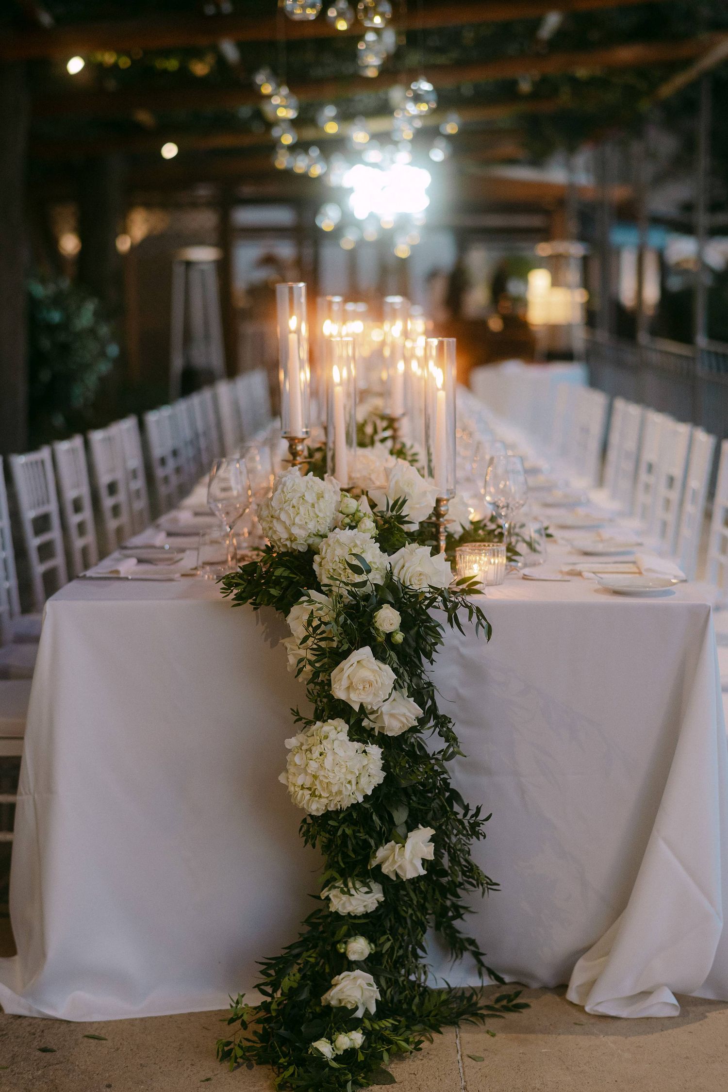Elegant wedding table with white linens, tall candles and cascading floral centerpiece of white roses and greenery.