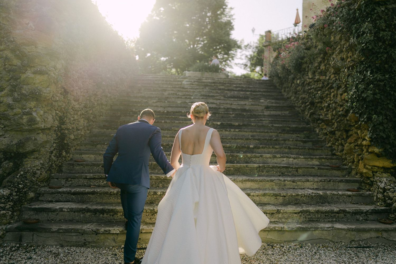 Newlyweds walk up stone steps surrounded by lush greenery as sunlight creates a dreamy backlit effect.