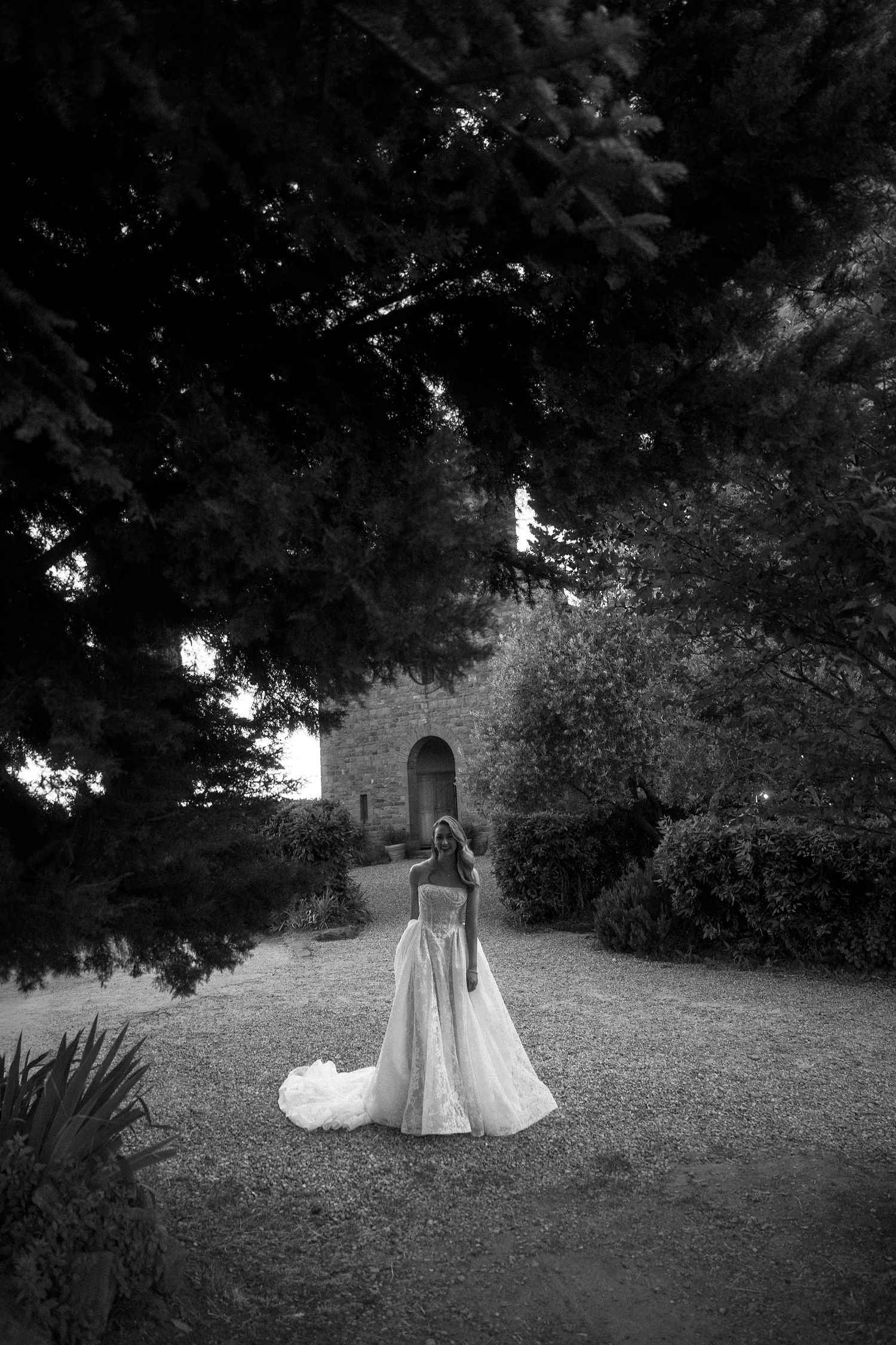 Black and white photo of a bride in a flowing gown standing on a gravel path surrounded by lush trees near a stone building.