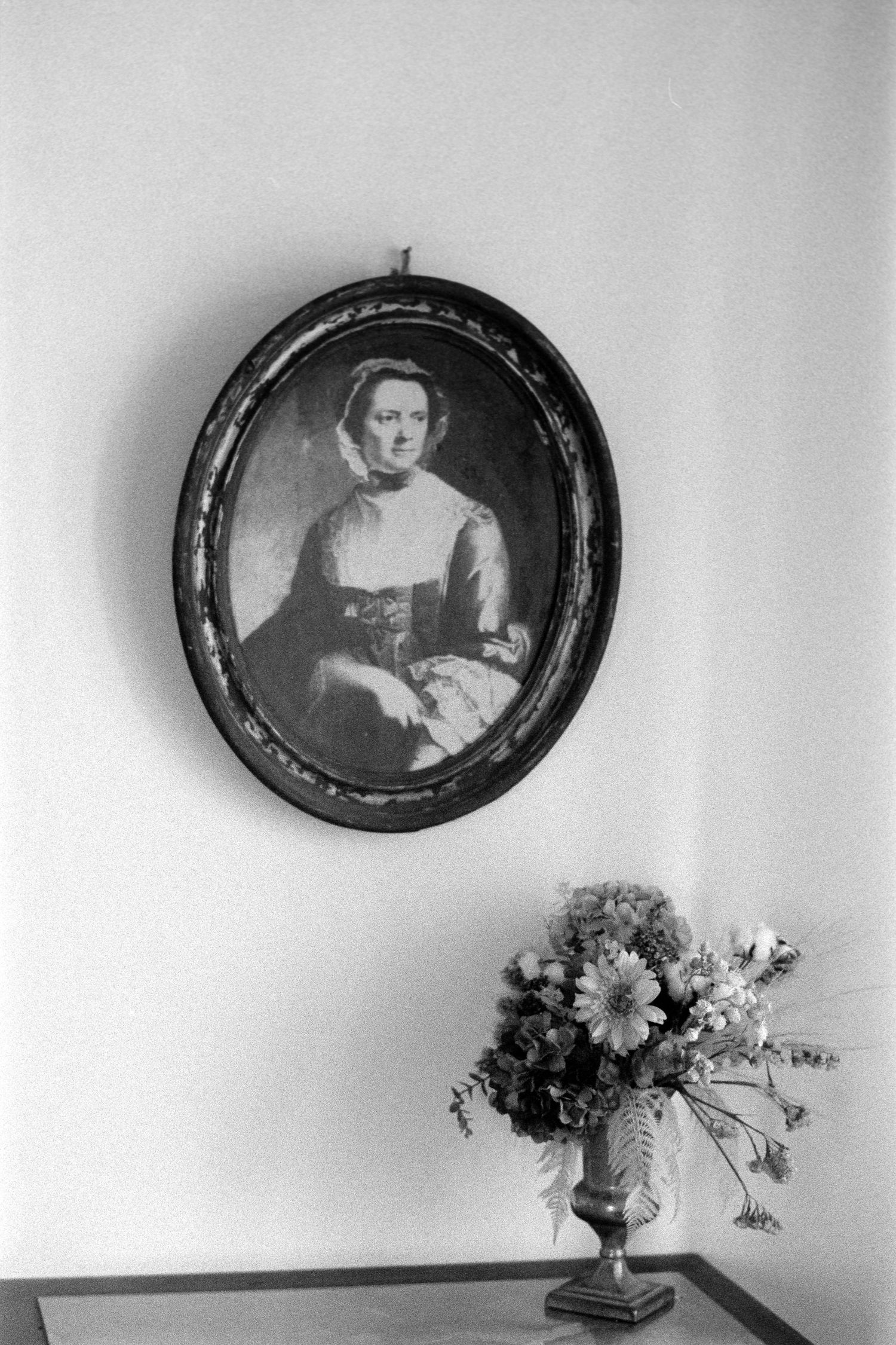 Black and white photograph of an antique oval portrait hanging above a floral arrangement in a vase.