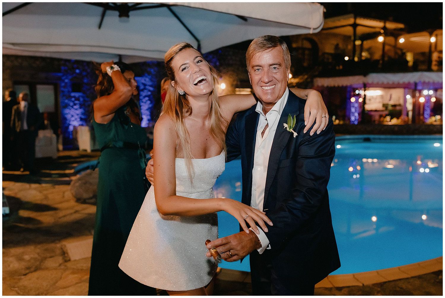 A joyful wedding celebration takes place outside at night by an illuminated swimming pool at a venue patio.