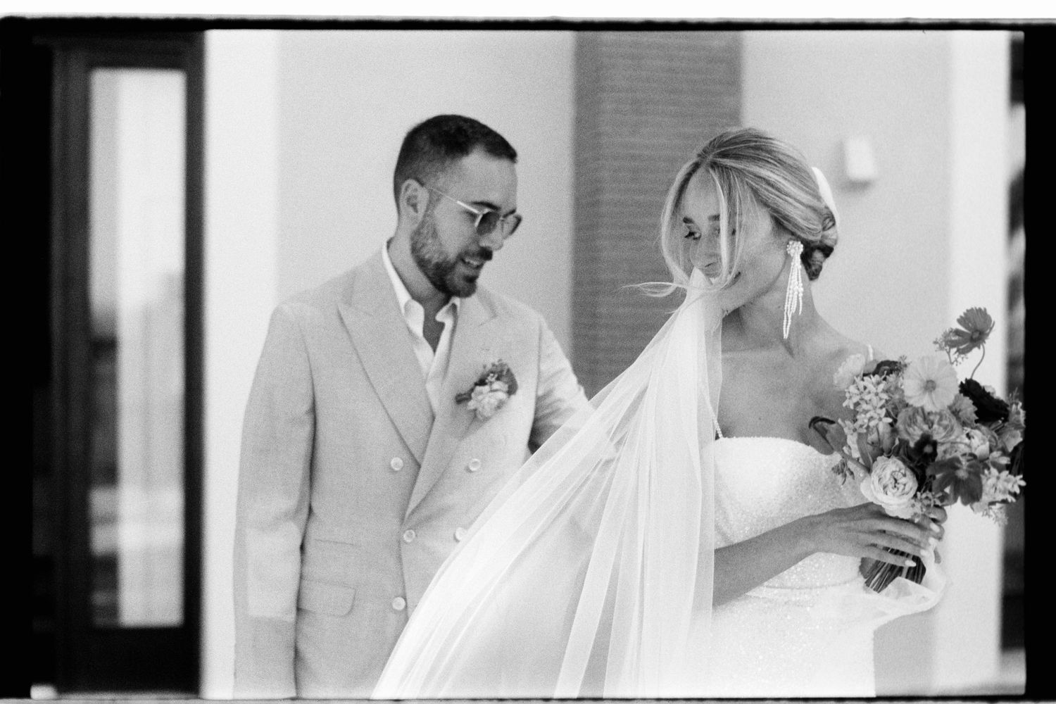 Black and white wedding photo showing bride in flowing veil with bouquet and groom in light colored suit and glasses.