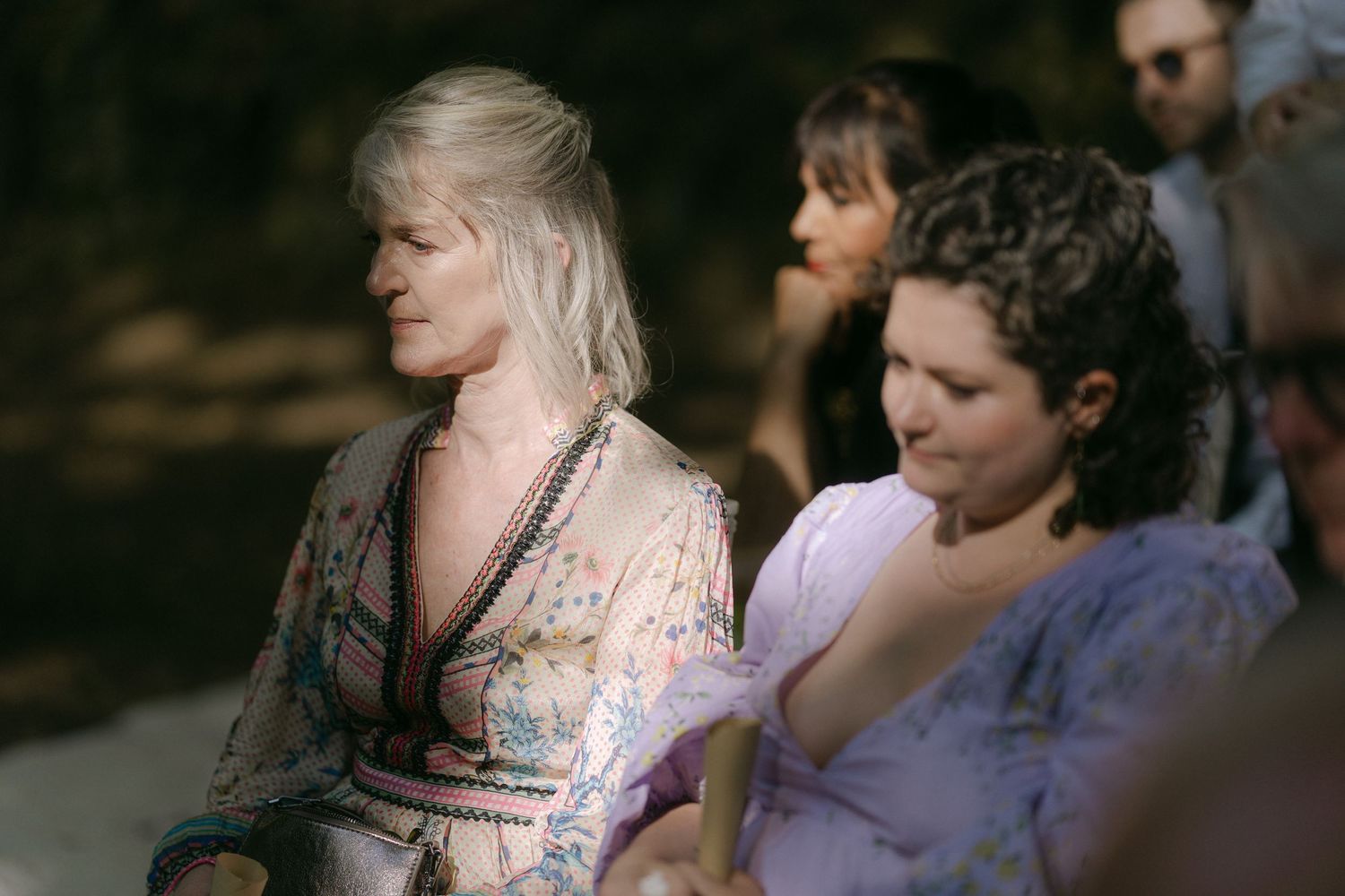 Two people sit together outdoors wearing vintage-style dresses in soft lighting.