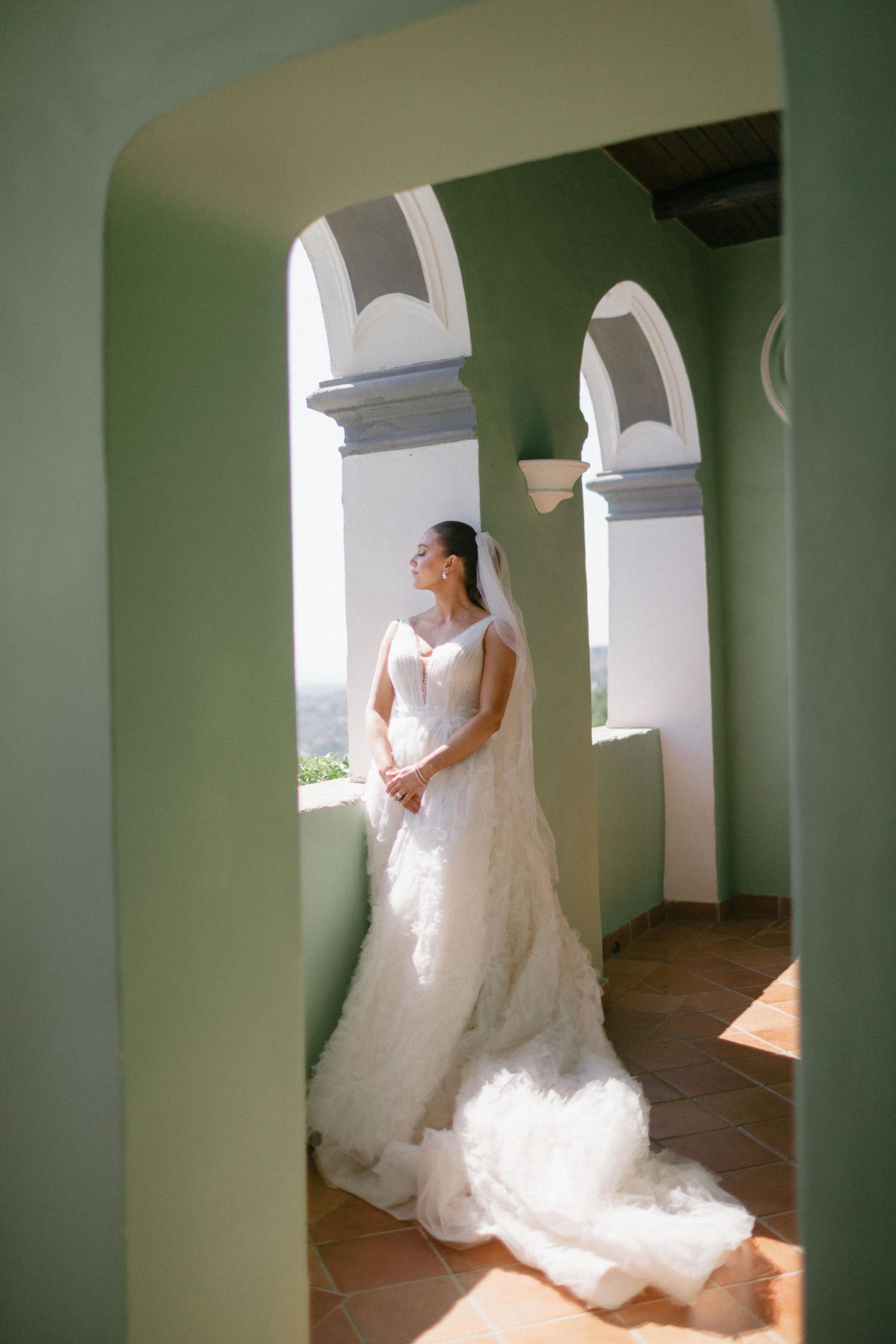 Sunlit bridal portrait showing an elegant white wedding gown with flowing train in an arched Mediterranean-style corridor.