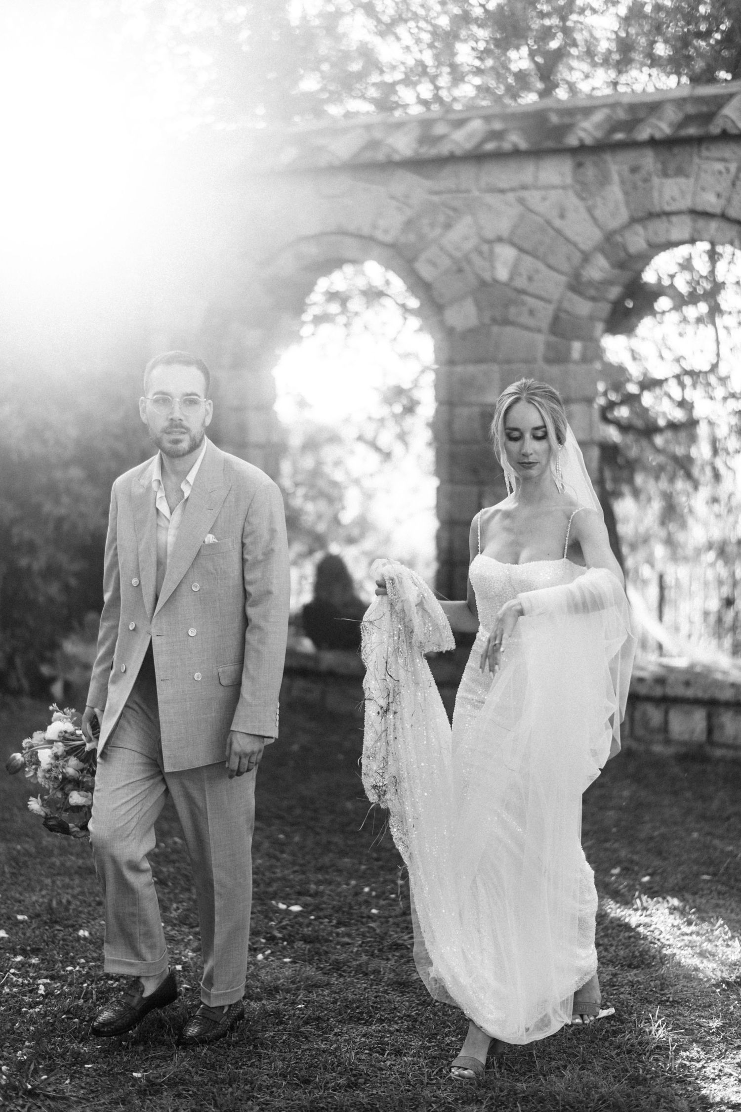 A romantic black and white wedding photo shows a couple walking together under an arched stone structure in soft sunlight.