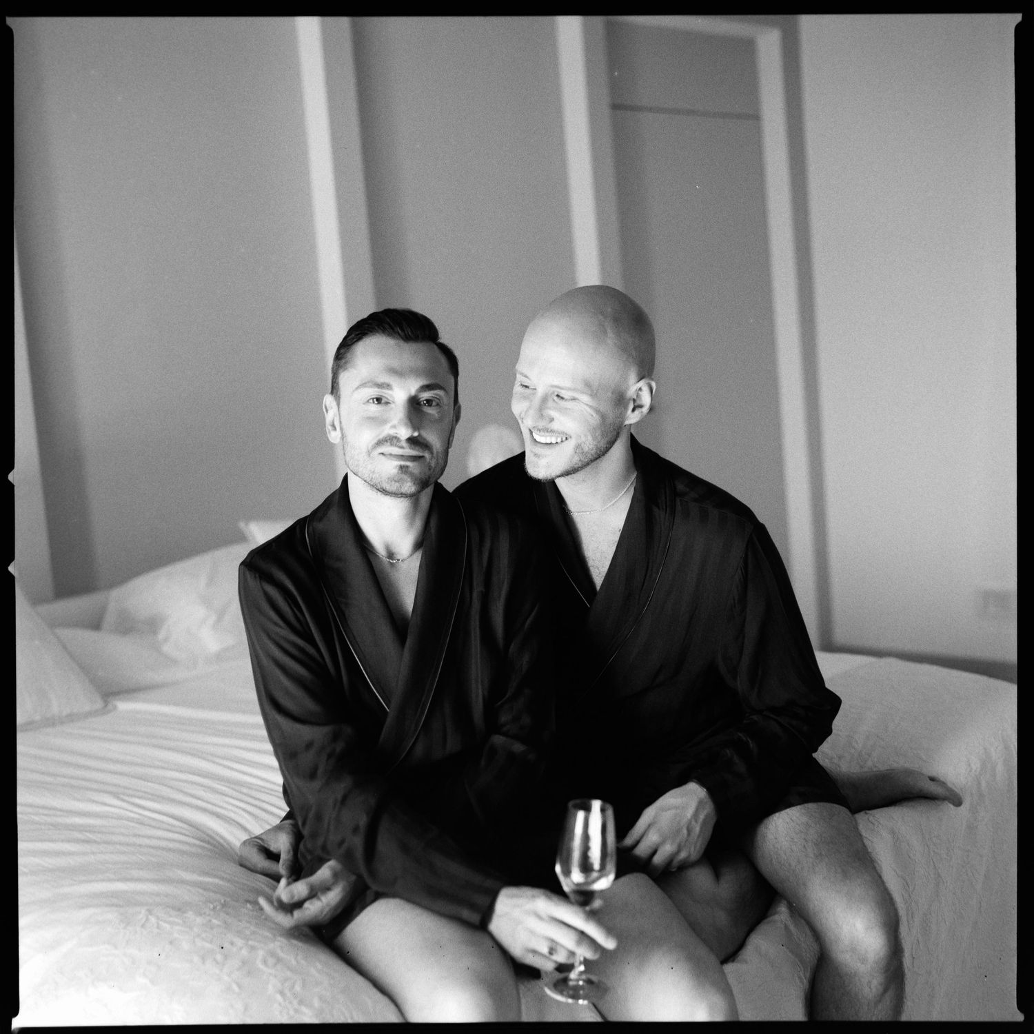 Two people in black robes sit together on a bed holding drinks in an intimate black and white portrait.