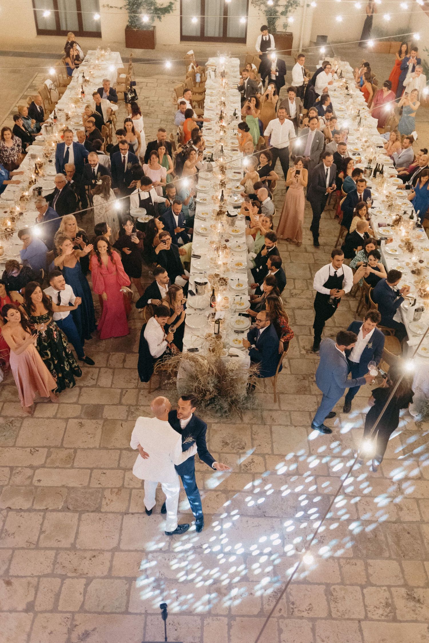 Overhead view of an elegant wedding reception with long banquet tables and couples dancing on a stone floor under string lights.