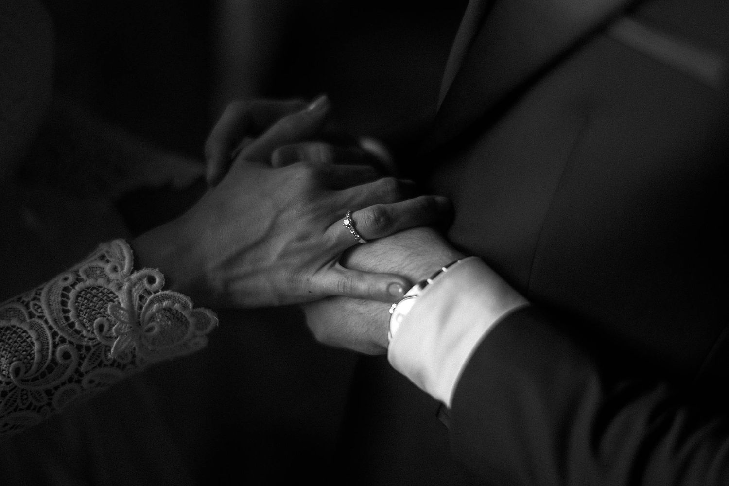 A black and white close-up of two hands holding each other, with one hand showing a detailed lace tattoo.