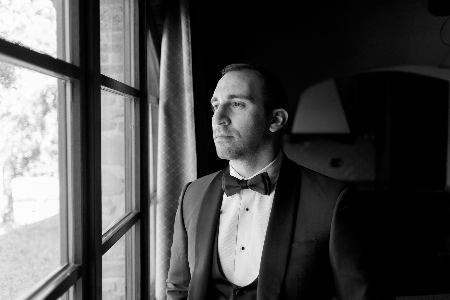 A person in a black tuxedo and bow tie stands near a window in a formal setting.