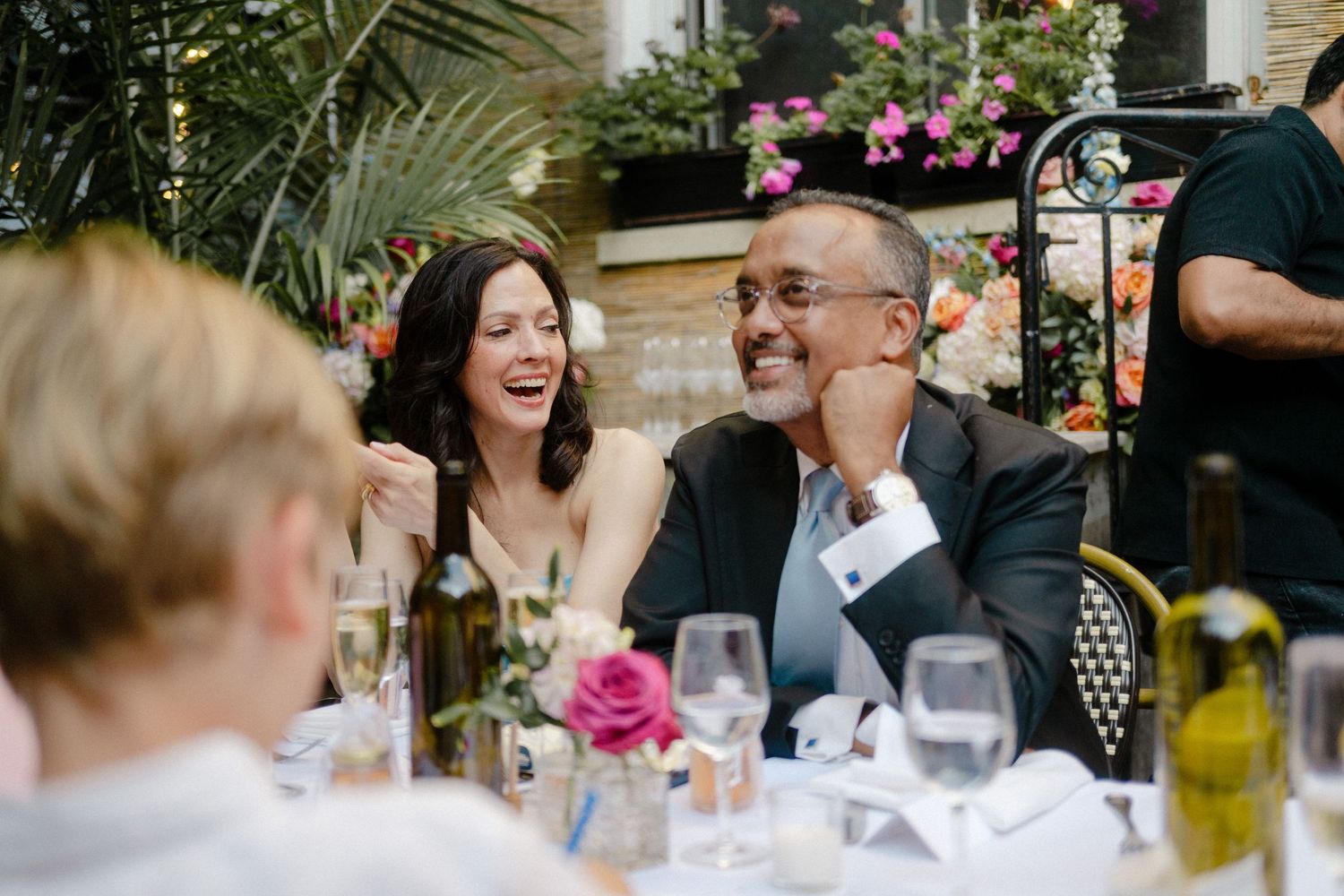 People share a joyful moment of laughter at an outdoor dining table with wine glasses and pink flowers.
