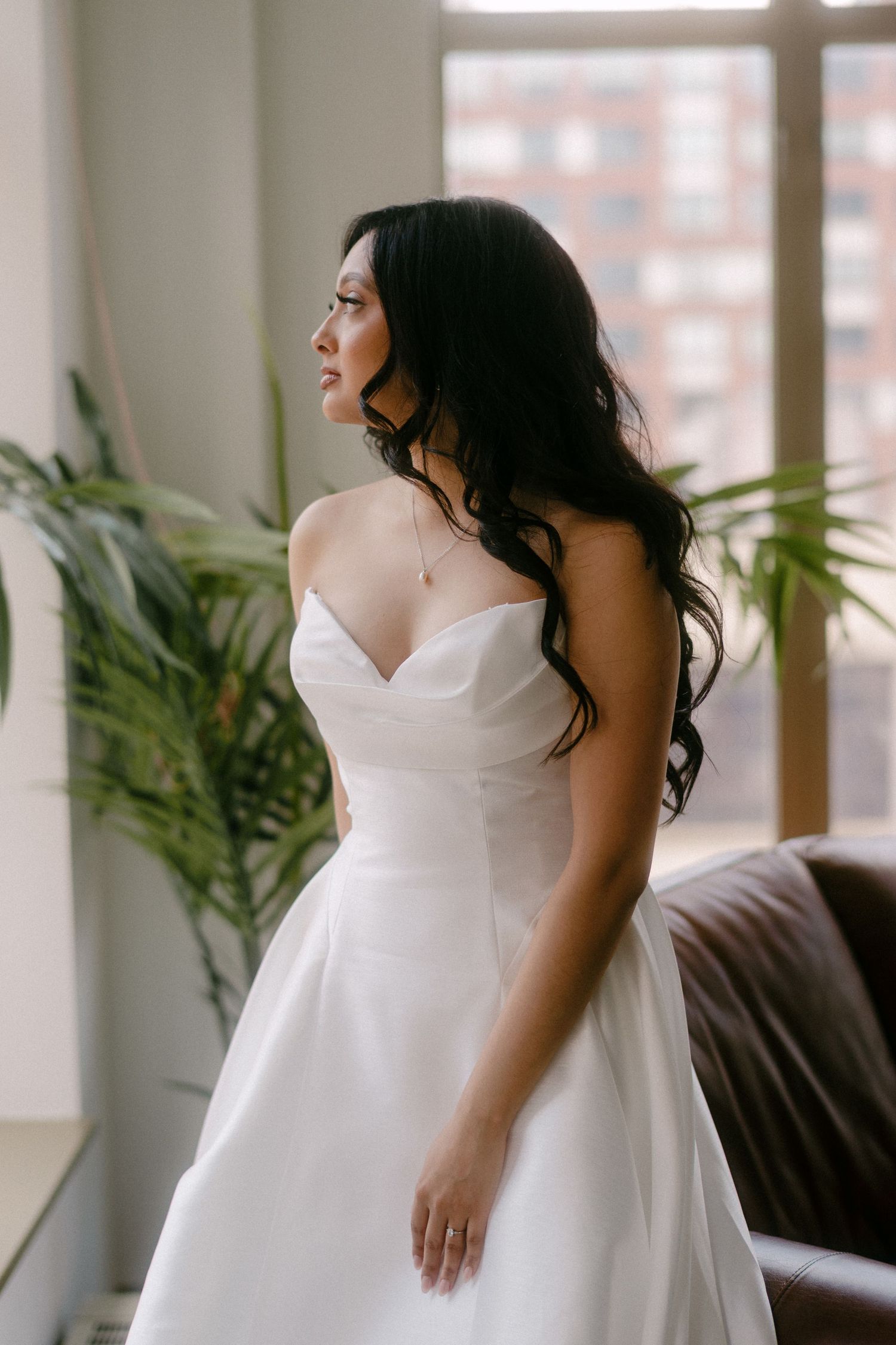 A bride in a white strapless wedding gown stands in a bright room with potted plants and windows in the background.