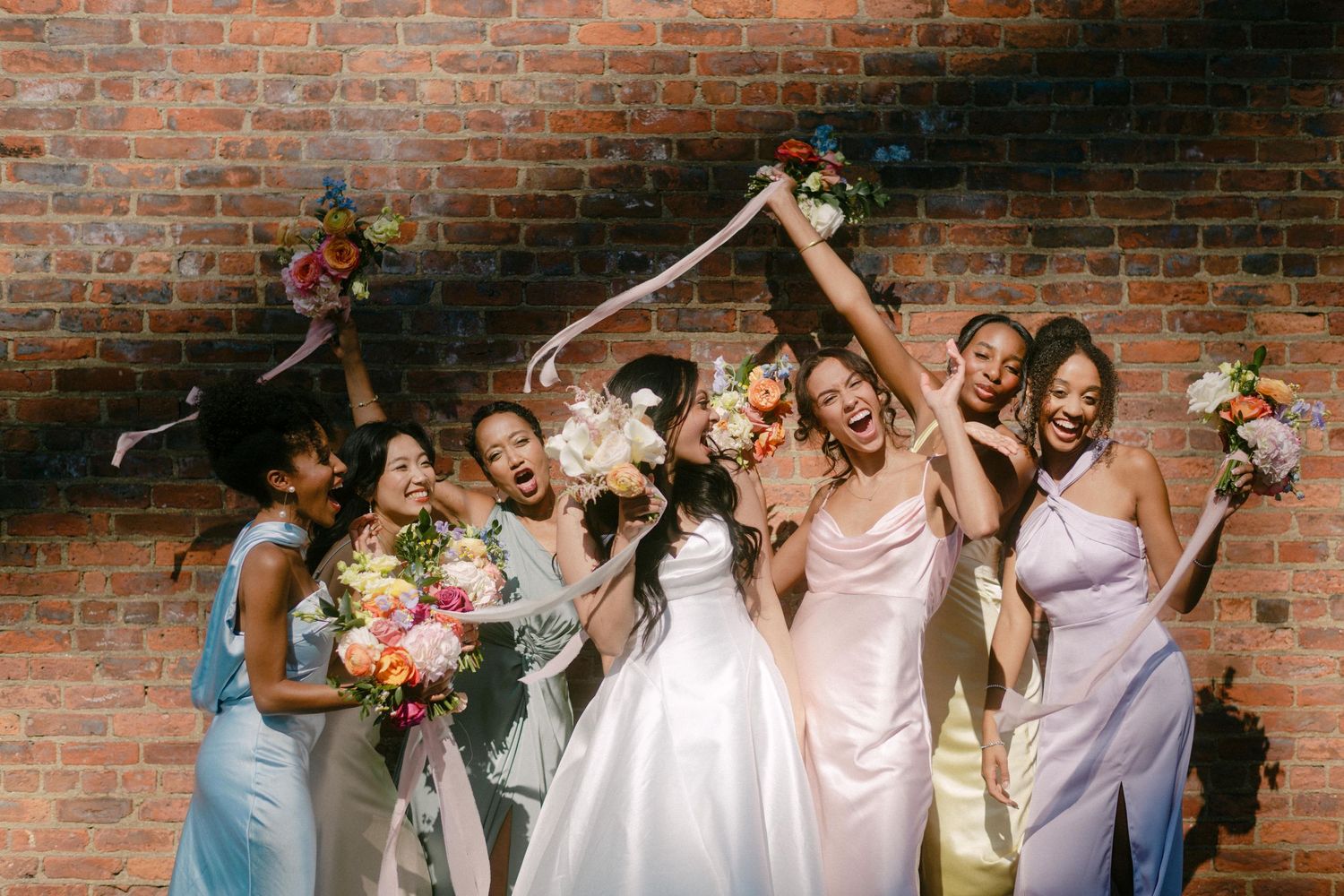 Wedding party celebrates joyfully in front of rustic brick wall with colorful floral bouquets.