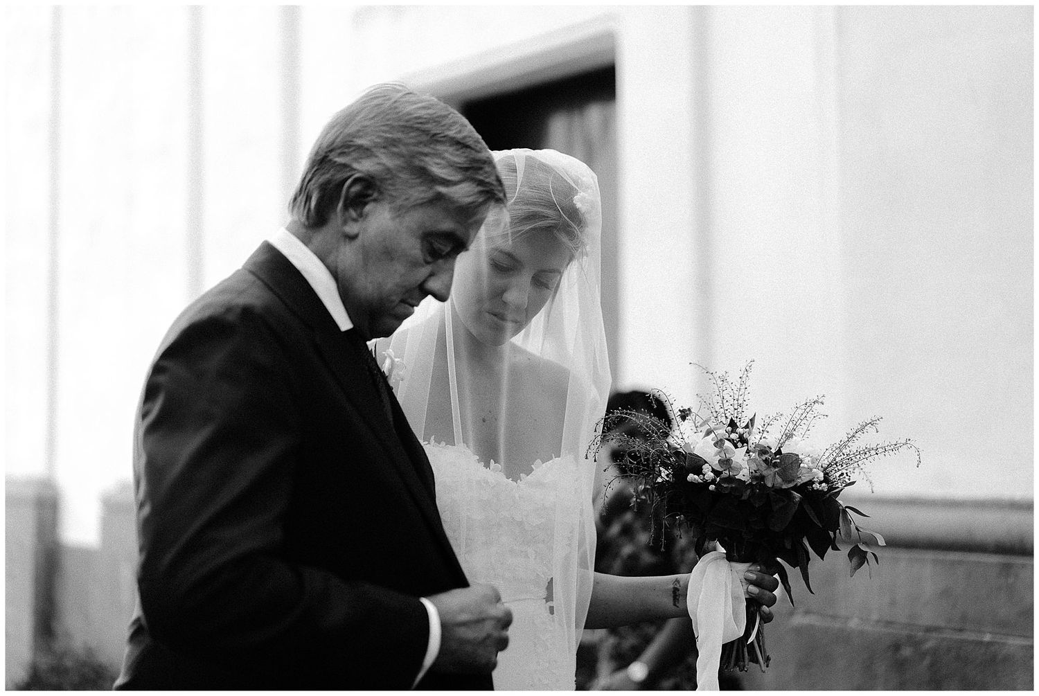 A black and white photograph shows a bride in a white veil next to someone in a dark suit holding a bouquet of flowers.