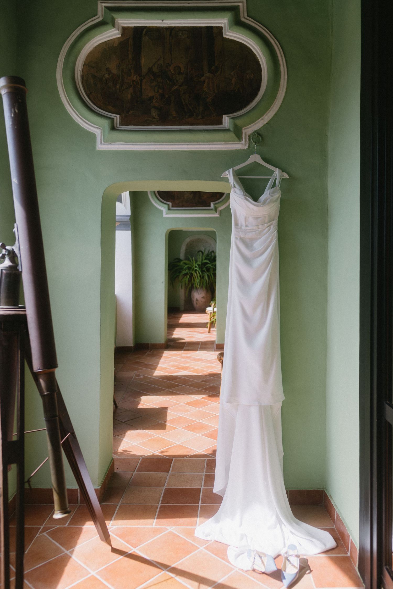 A white wedding dress hangs in a green-walled hallway with terracotta floors and natural lighting streaming through.