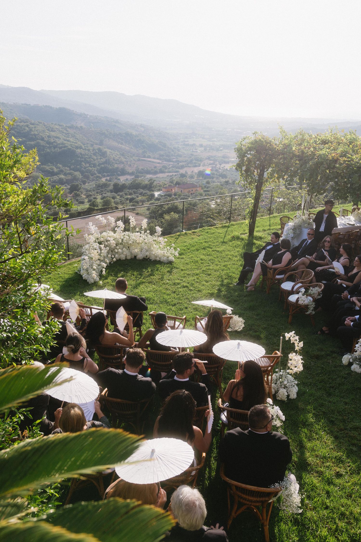 Guests seated at round tables on a scenic hillside terrace overlooking a lush valley during an outdoor event.