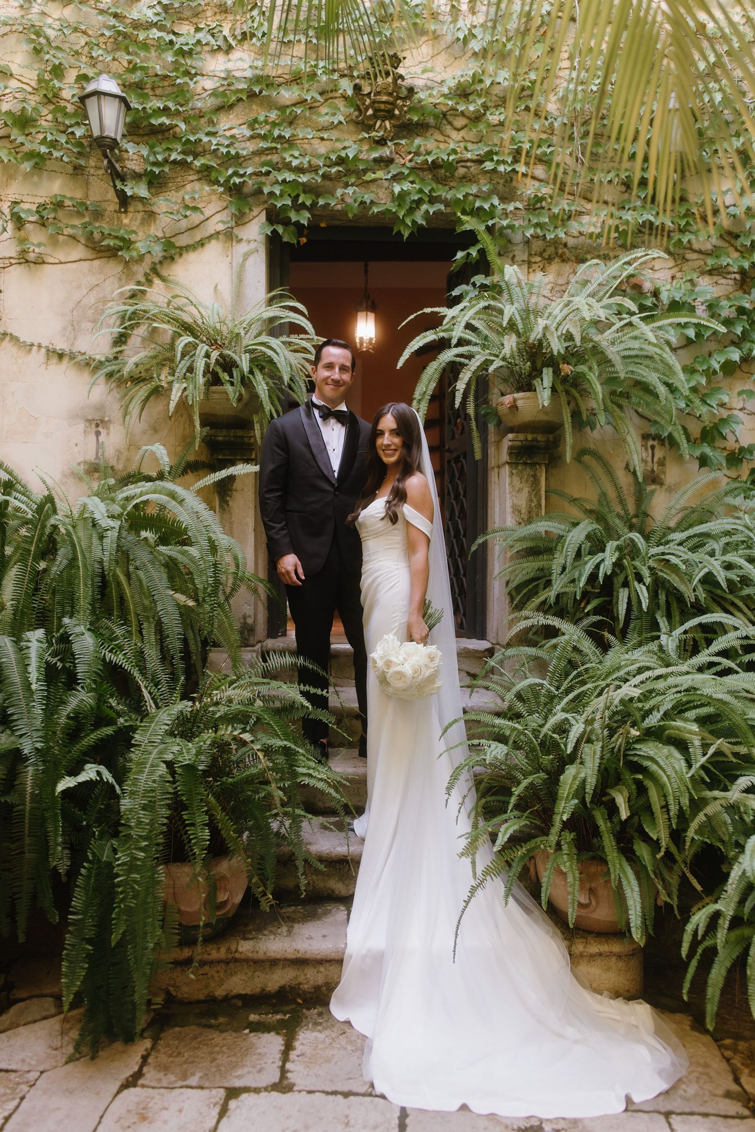 A romantic wedding photoshoot in a lush garden courtyard with ferns and palm fronds creating a tropical atmosphere.